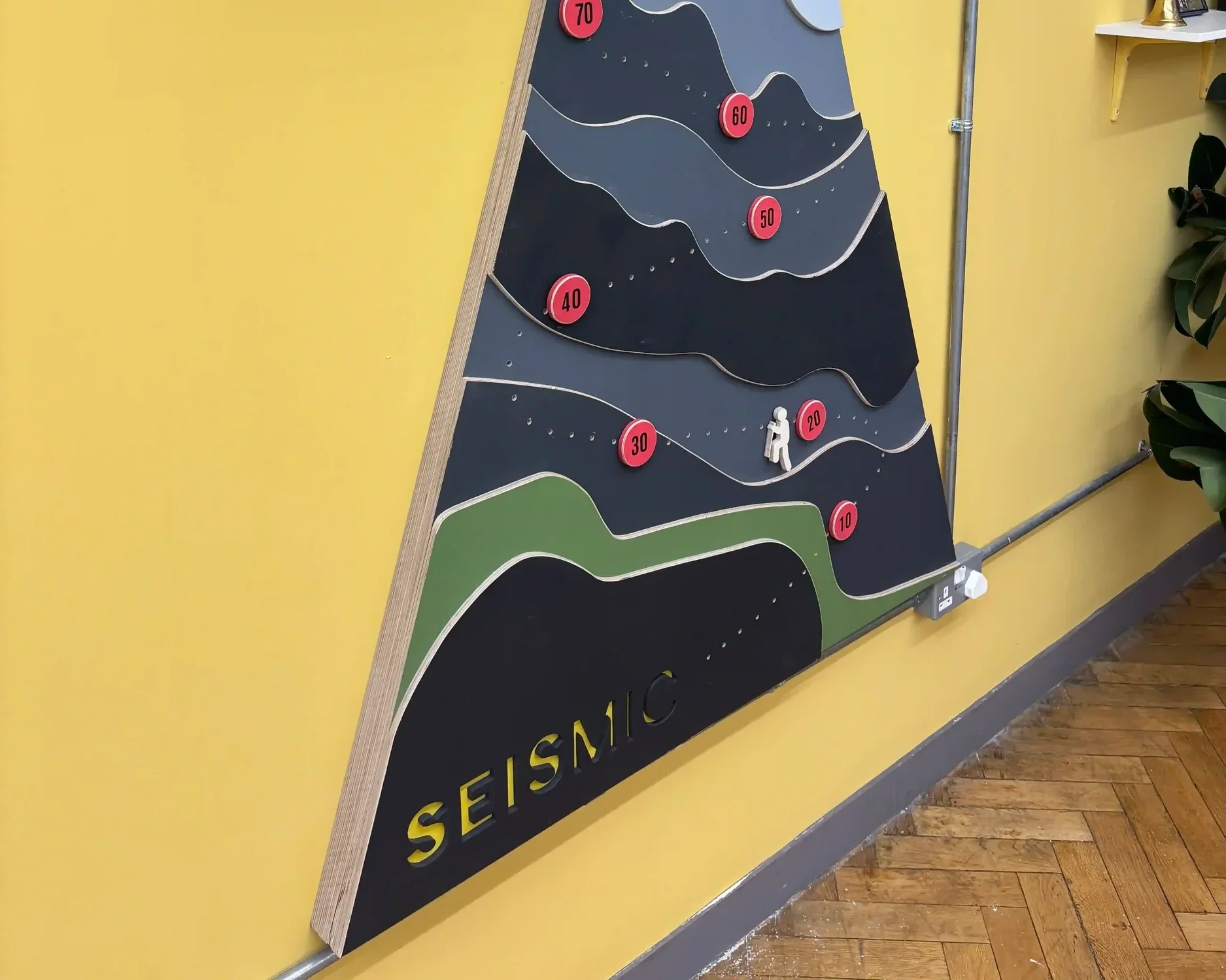Seismic bespoke branded triangular interactive display board with mountain graphic on yellow office floor surrounded by plants