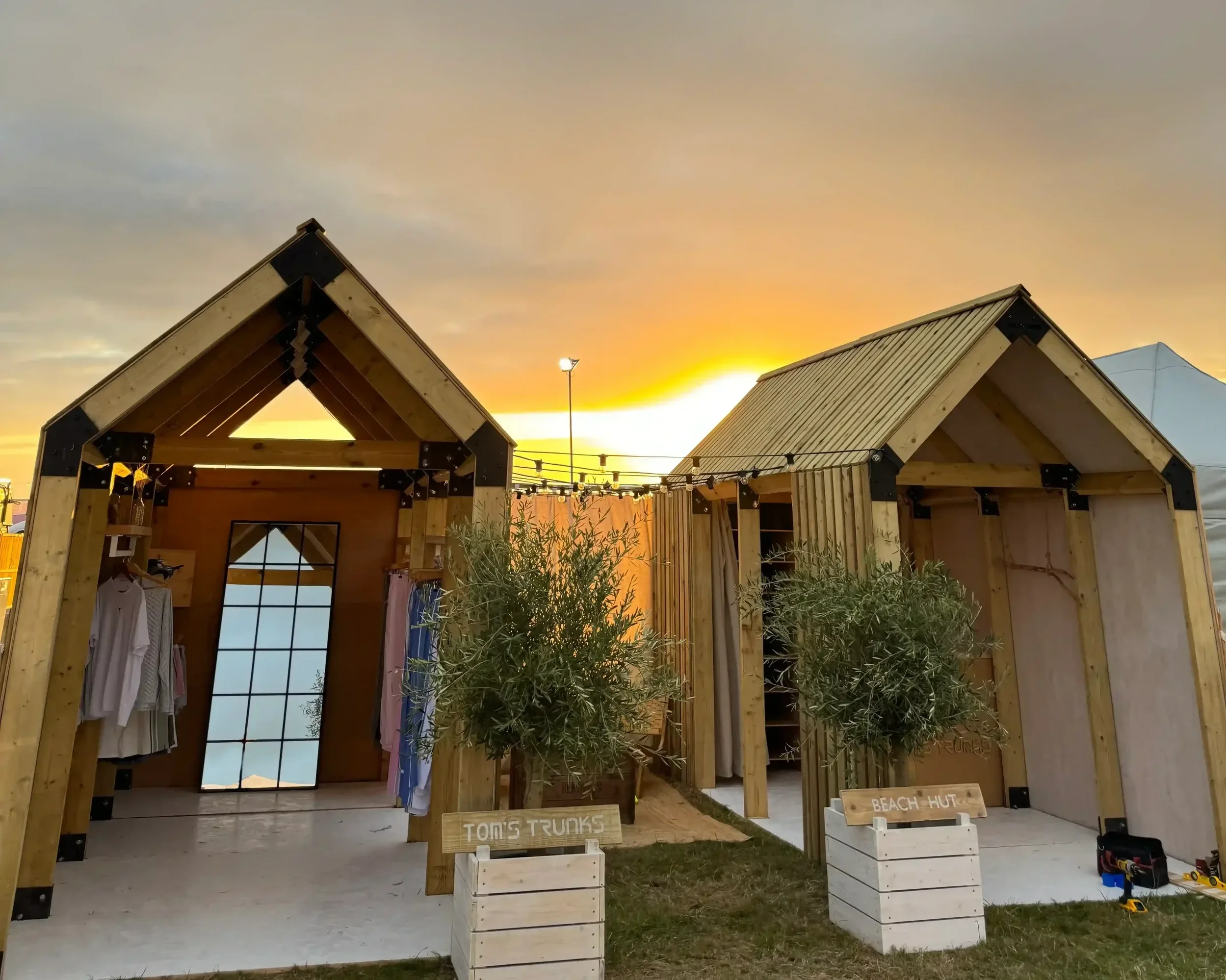 Tom's Trunks bespoke wooden beach hut brand activation structures at outdoor festival during golden sunset with potted olive trees