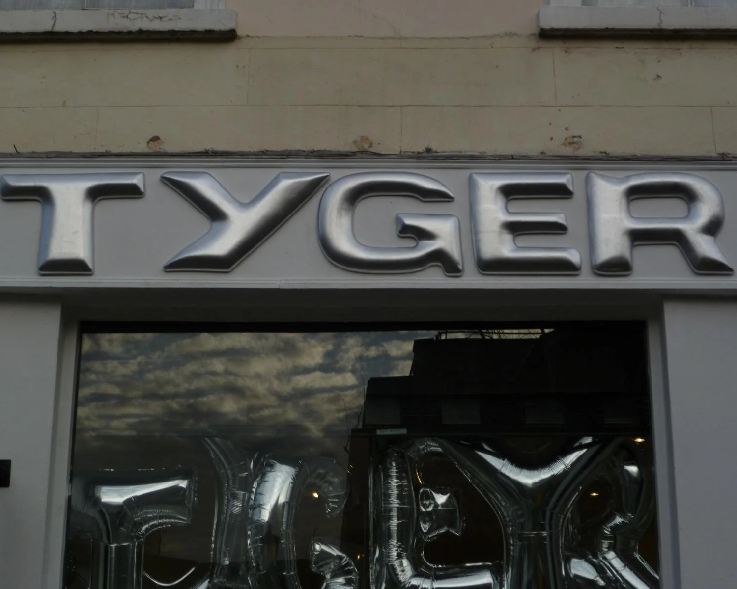 Close-up of a metallic sign reading 'TYGER' above a window reflecting clouds and a building.