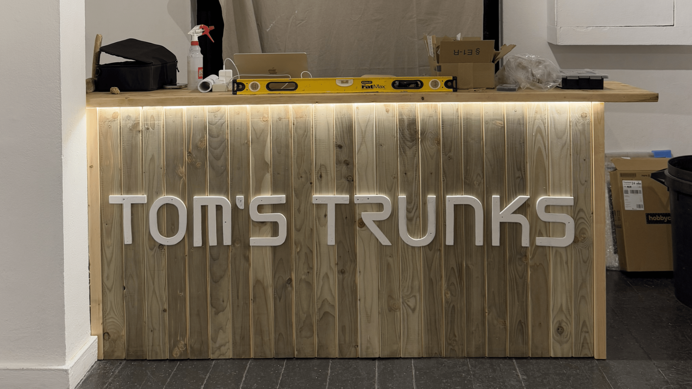Wooden bar with white illuminated sign reading 'TOIN'S TRINKS' in front of a work area with tools, boxes, and supplies.