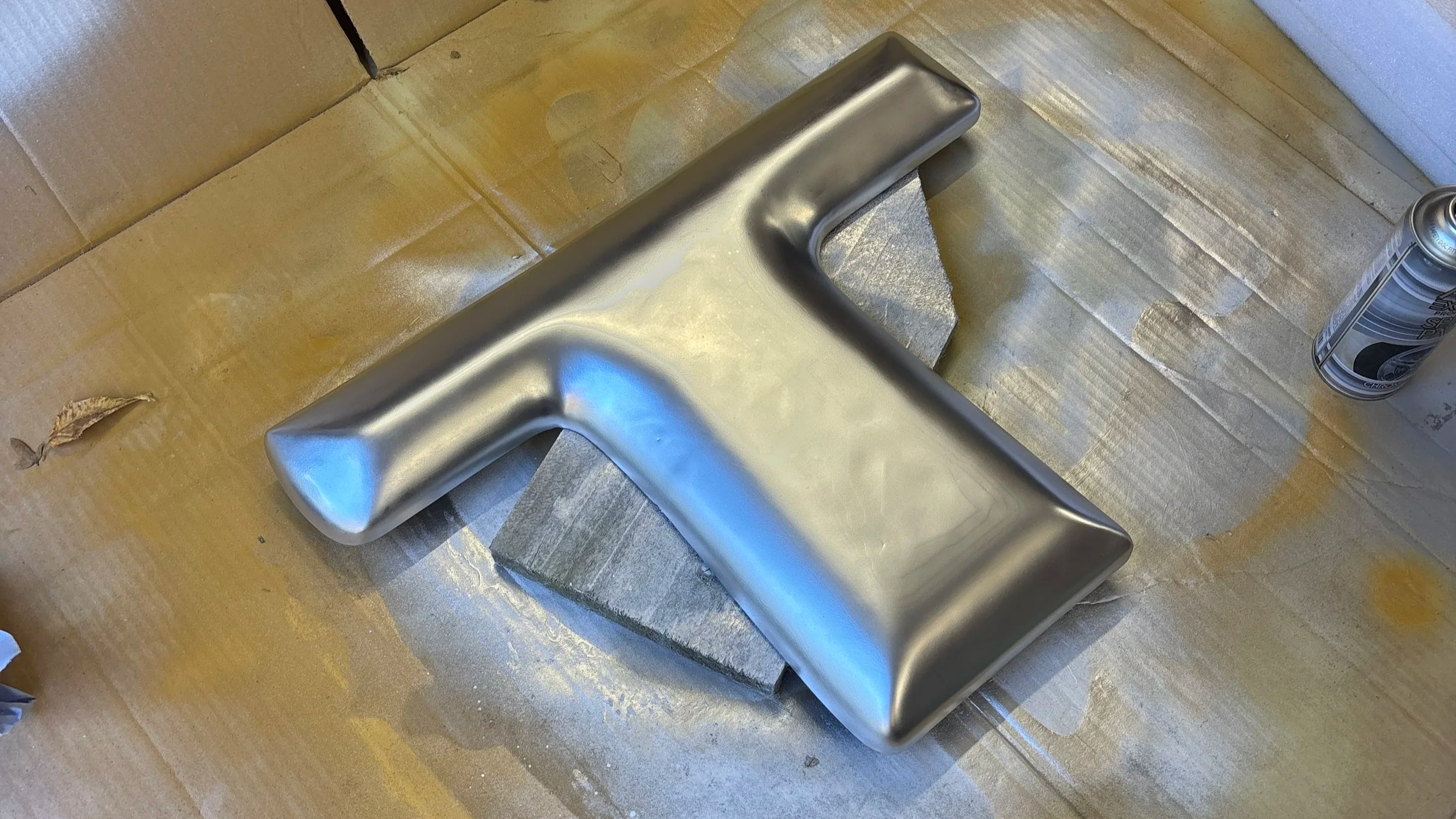 A metallic T-shaped object, possibly a sculpture or a tool, resting on a piece of wood with a can of spray paint nearby and a cardboard background.