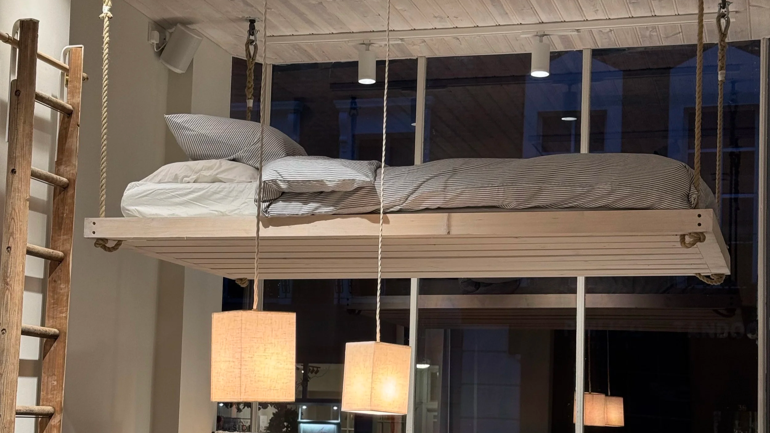 A wooden swing bed with striped bedding and pillows hanging from a exposed ceiling in a modern interior space with large glass windows and hanging lamps.