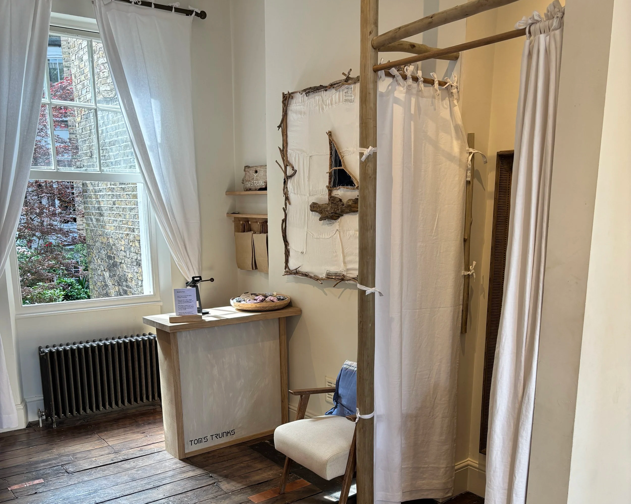 A room with a large window, white curtains, a wooden mannequin, and a display of fabric or textile pieces on a wall behind the mannequin. There is also a small table with objects, a radiator under the window, and a wooden floor.