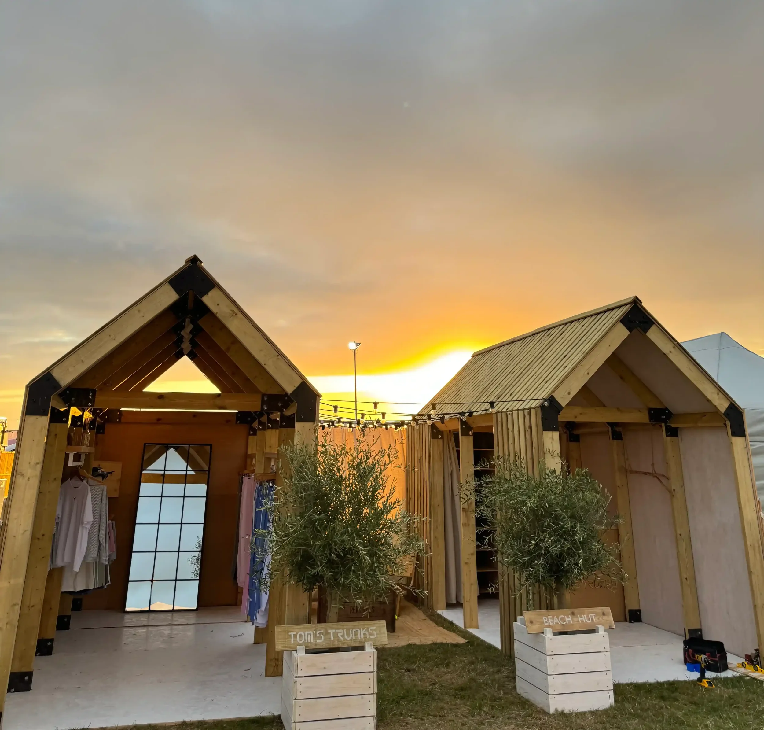 Tom's Trunks bespoke wooden beach hut brand activation structures at outdoor festival during golden sunset with potted olive trees
