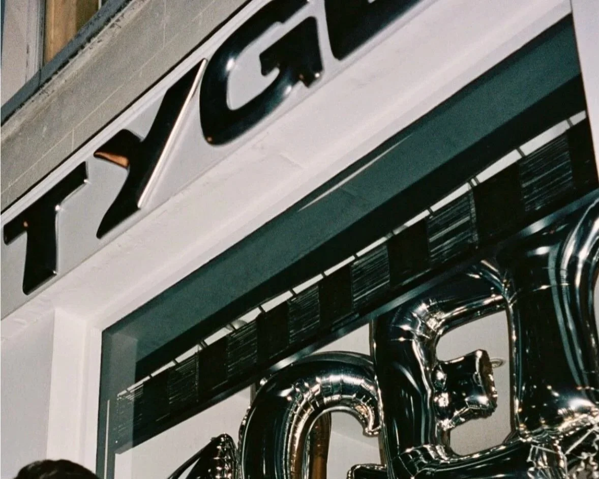 Close-up of a sign with metallic letters spelling "YEC" above a window with black and white striped decorations; silver balloons are partially visible at the bottom right.