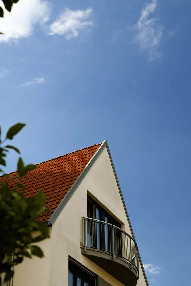 A modern white house with a sloped red-tile roof and a curved balcony, against a bright blue sky with a few clouds.