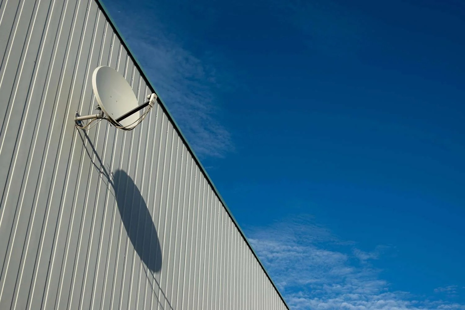 Satellite dish mounted on the side of a building with a shadow cast on the wall, against a partly cloudy blue sky.