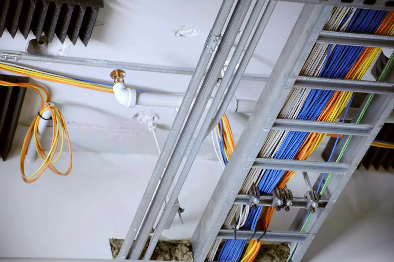Electrical and data cables organized and routed through a metal cable tray and conduit, with a ladder leaning against the wall in an unfinished or under-construction room.