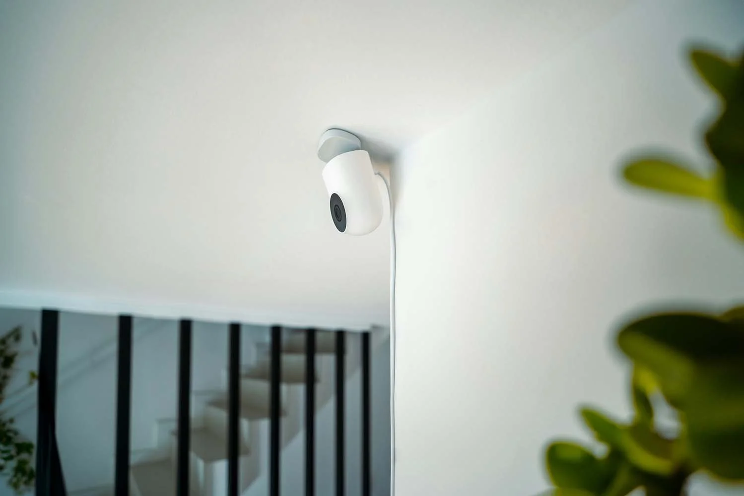 A white security camera mounted on a ceiling, with a black lens, and a cord hanging down.