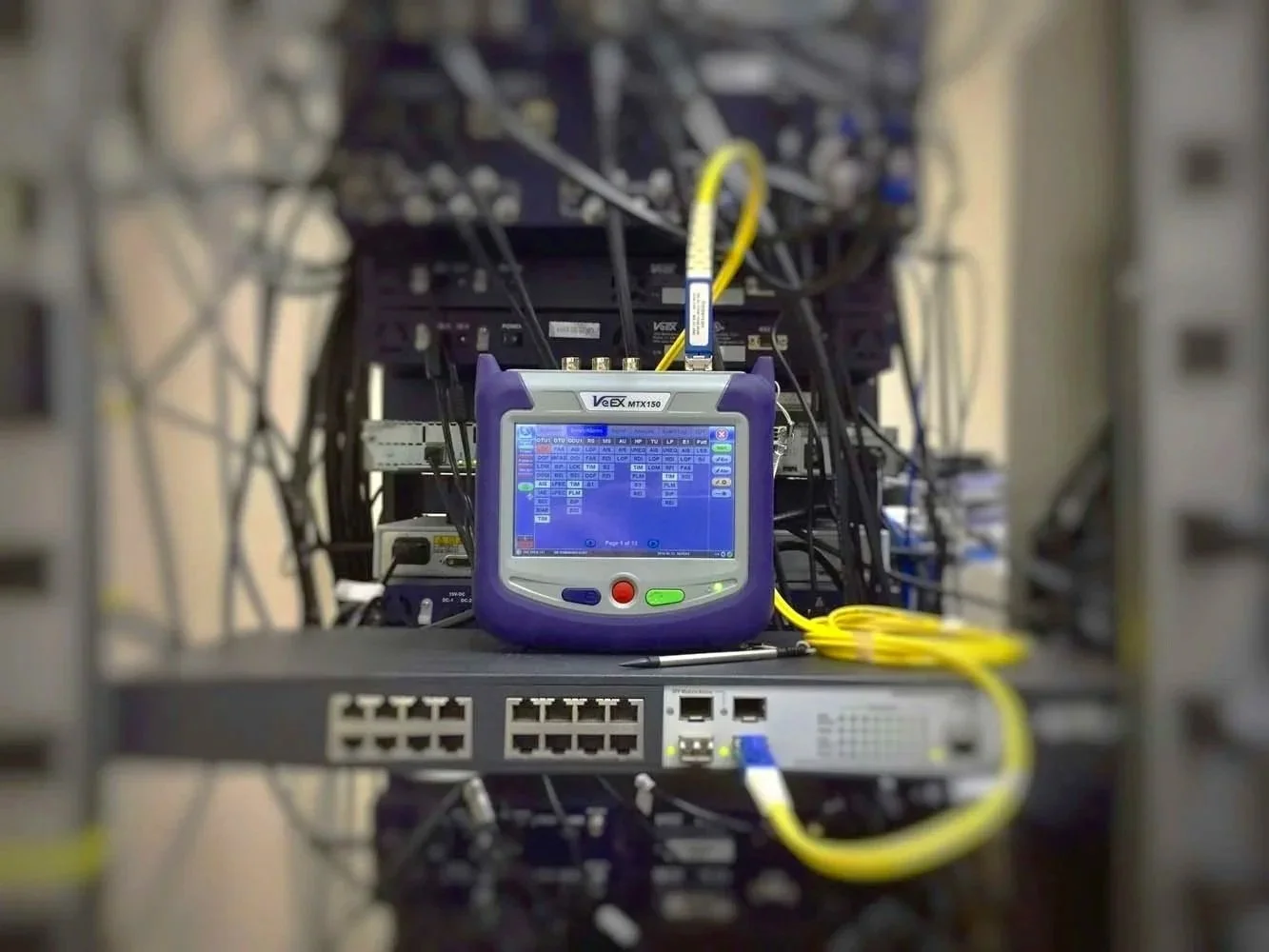 Close-up of a fiber optic network testing device on a server rack, with multiple Ethernet cables connected to networking equipment.