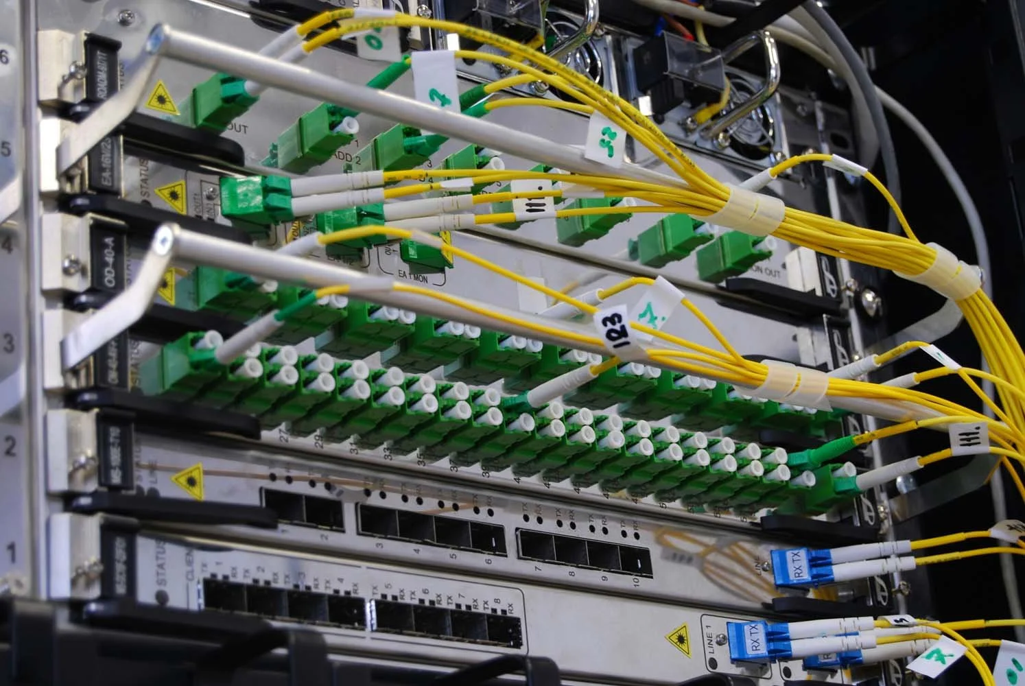 Close-up of a fiber optic patch panel with yellow and white cables connected to green adapters, with labels and switches on a metallic rack.