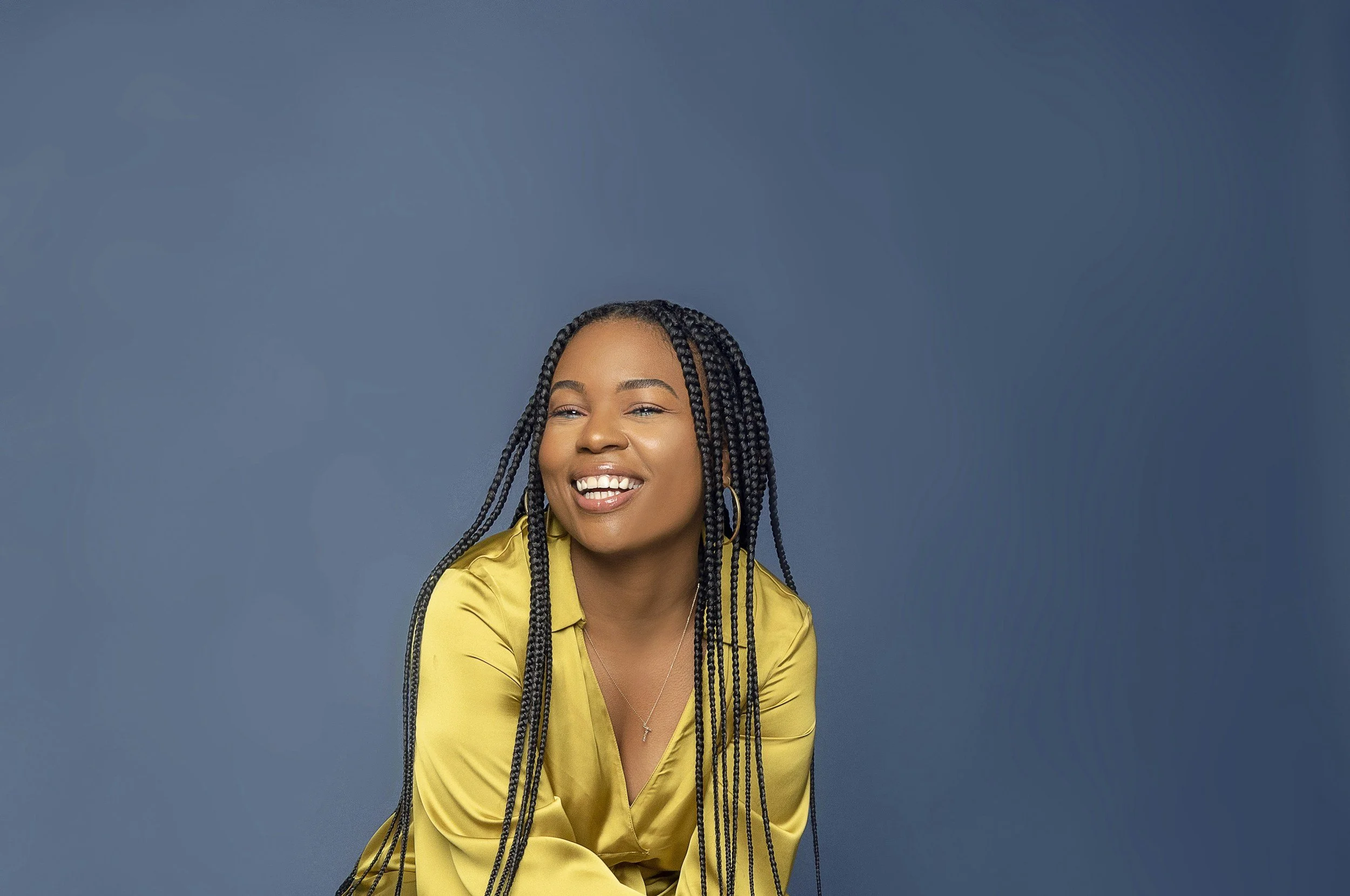 A smiling female counsellor with long braids wearing a yellow satin shirt against a blue background.