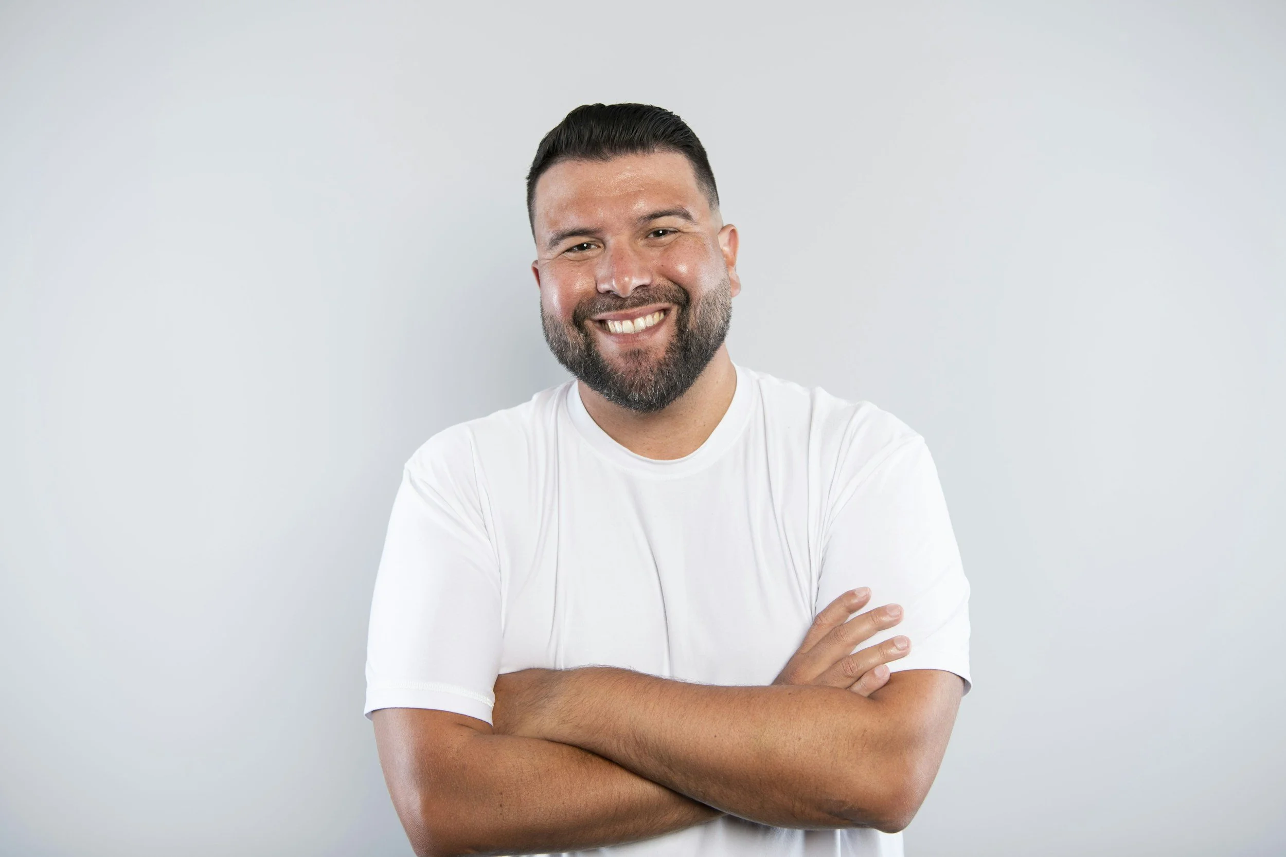Smiling man with a beard and short dark hair, wearing a white t-shirt, standing against a plain light gray background.
