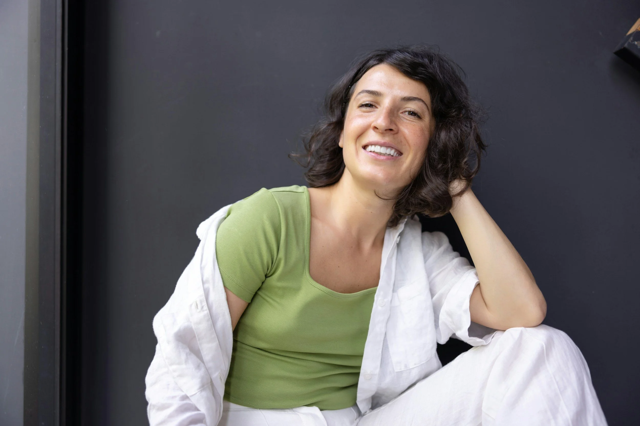 A female psychotherapist with short dark hair and freckles smiling with her head tilted, wearing a light green top and white shirt, sitting against a dark background.