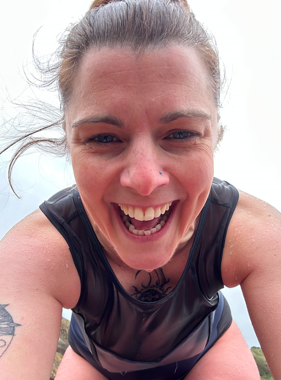 Close-up of a smiling woman with a nose piercing, tattoos on her arms, wet skin, wearing a black sleeveless top outdoors.