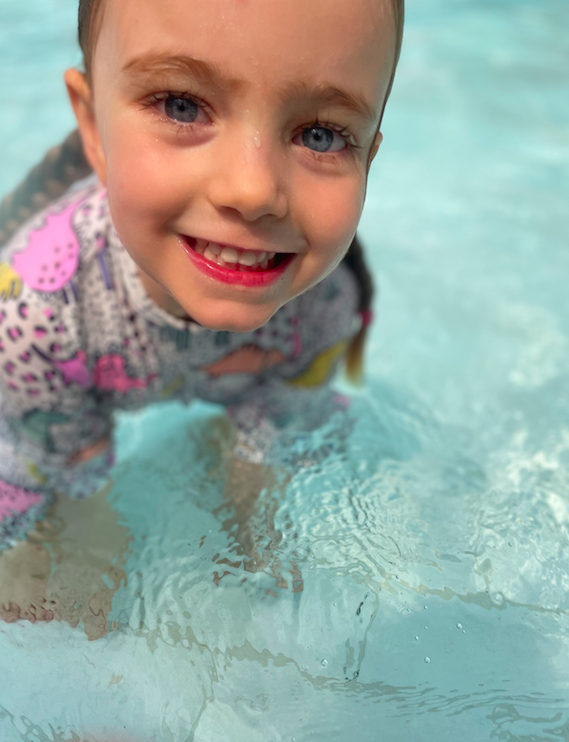 A smiling young girl with wet hair and blue eyes, wearing a colorful patterned swimsuit, in a swimming pool.