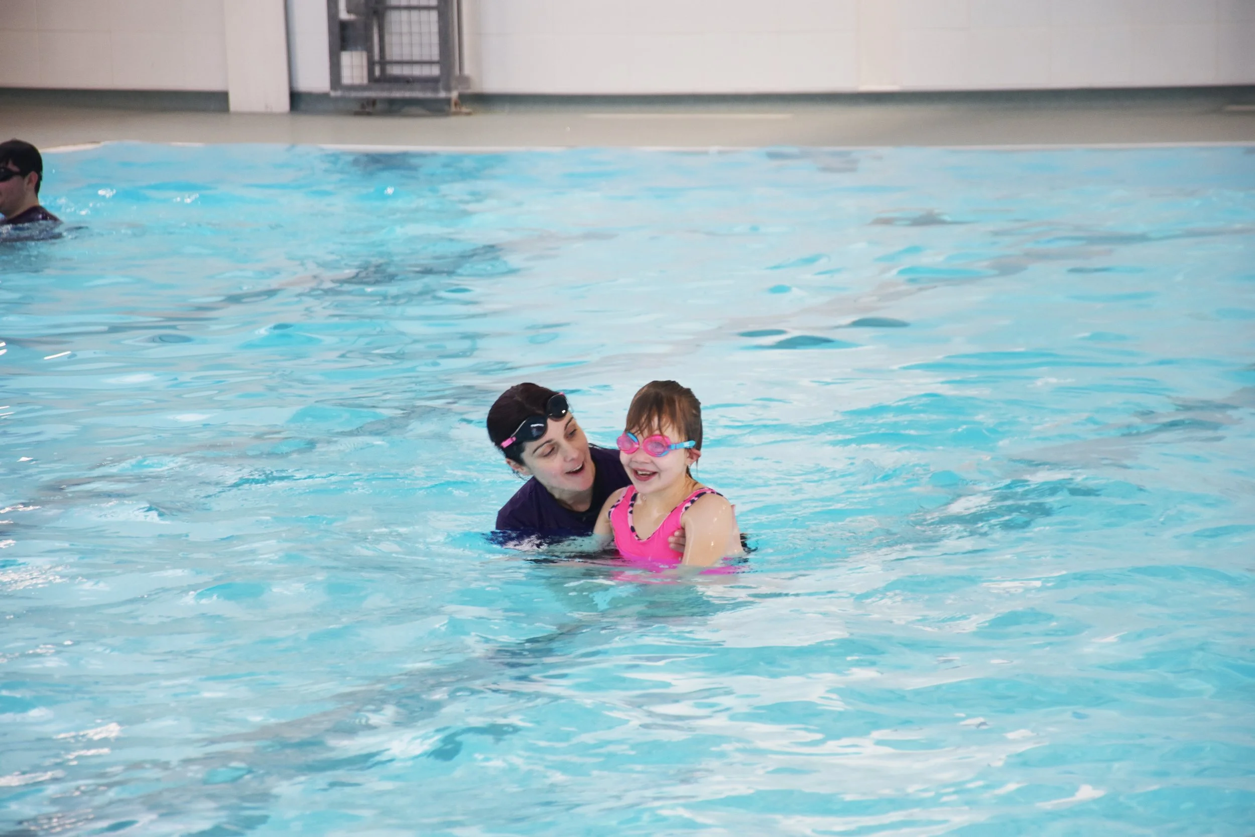 A young girl in a pink swimsuit and pink goggles smiling and enjoying swimming in a pool while being supported by an adult woman.