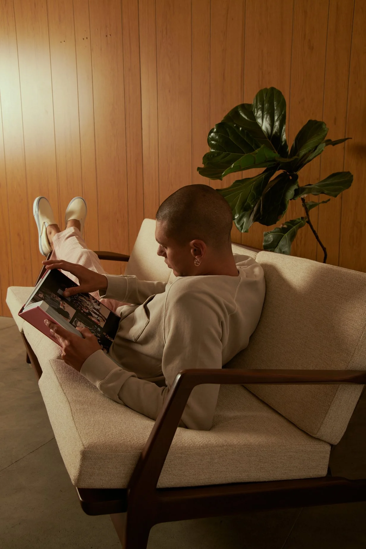 A young man with a shaved head and earrings is sitting on a beige couch, reading a photo album, with his feet resting on the armrest. He is in a room with wooden panel walls and a large leafy plant behind him.