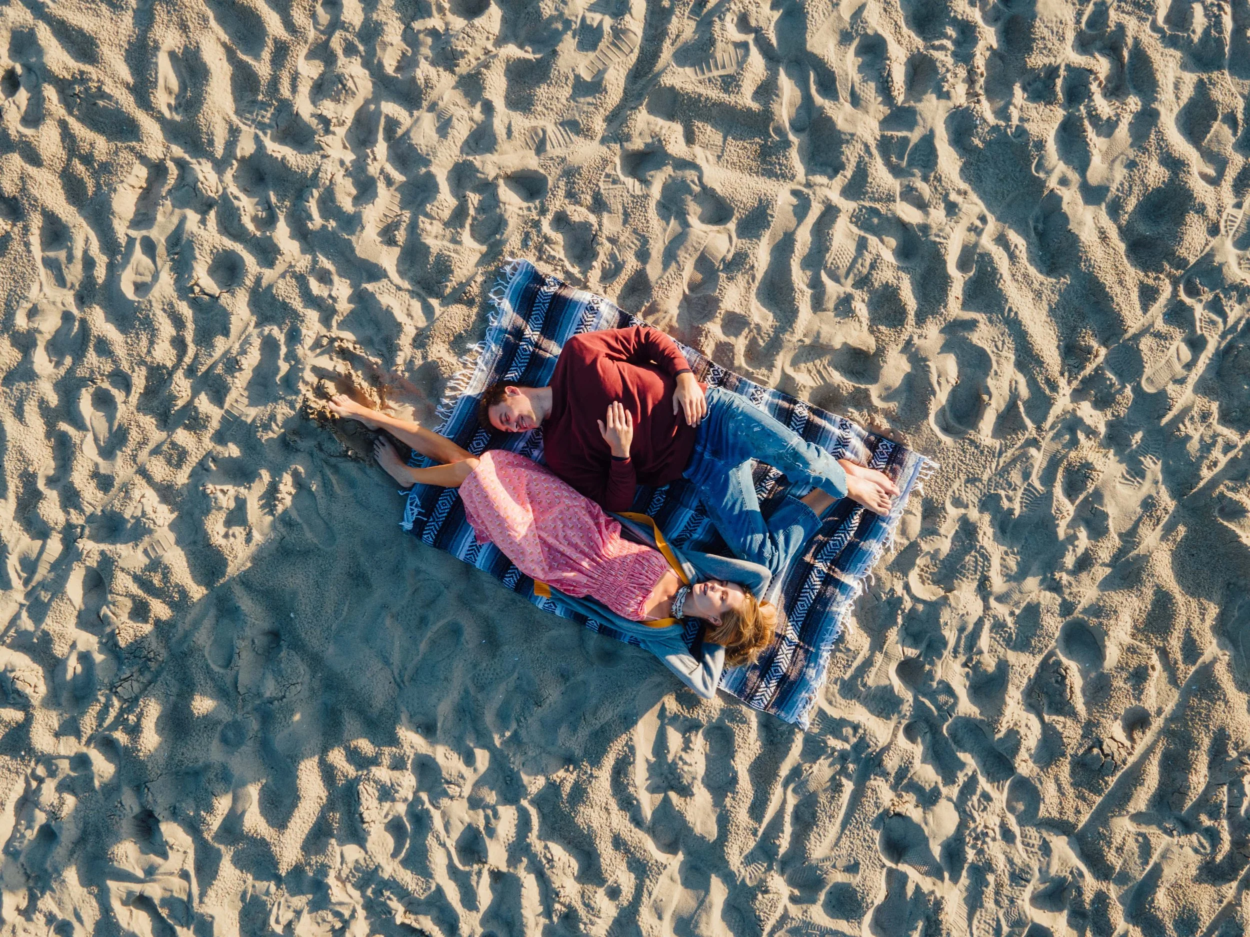 A woman and a girl lying on a striped blanket on sandy beach, with woman appearing to be relaxing with eyes closed and hand on chest, and girl resting with head on blanket.