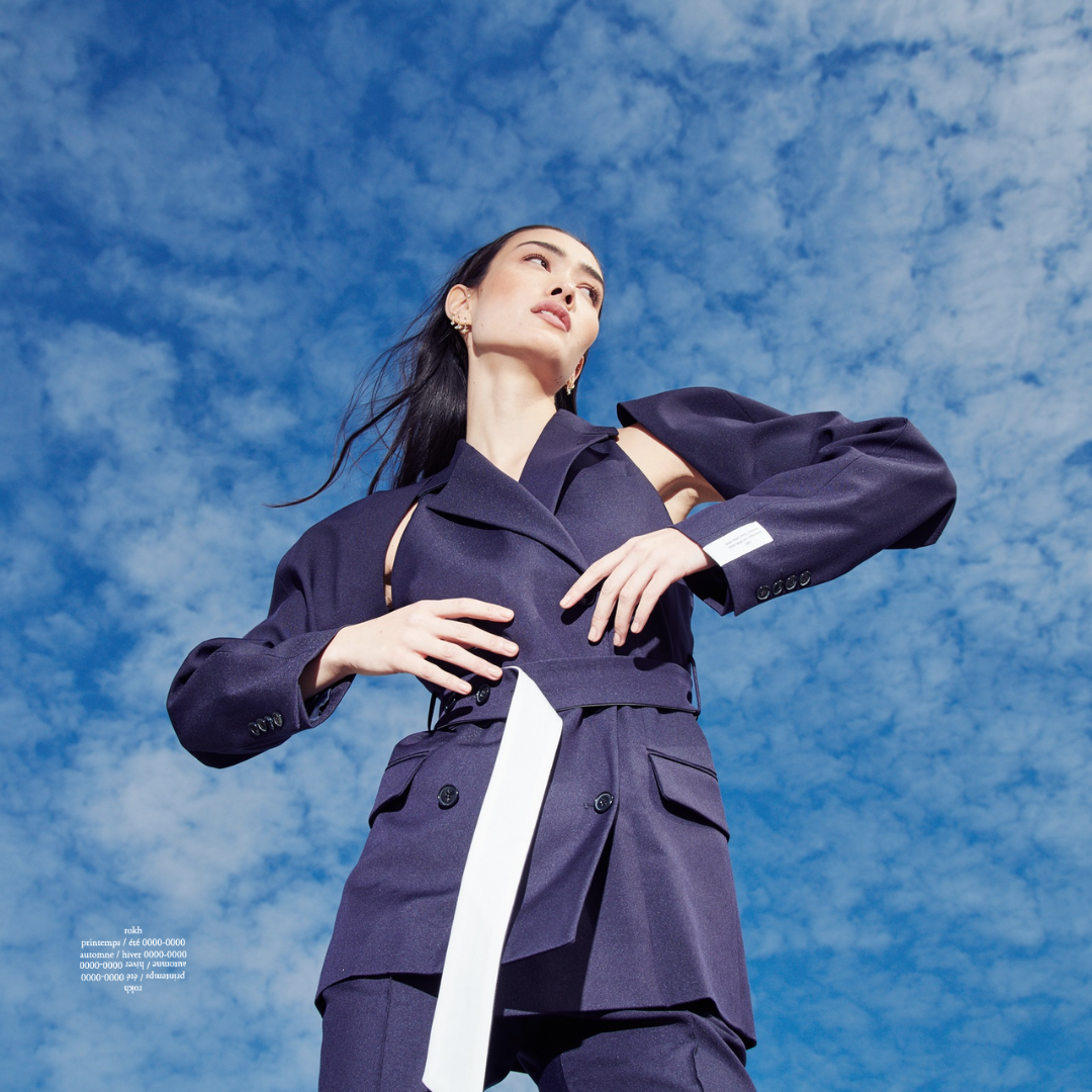 A woman in a dark navy fashion outfit standing outdoors under a partly cloudy sky, looking upward with her hands on her waist.