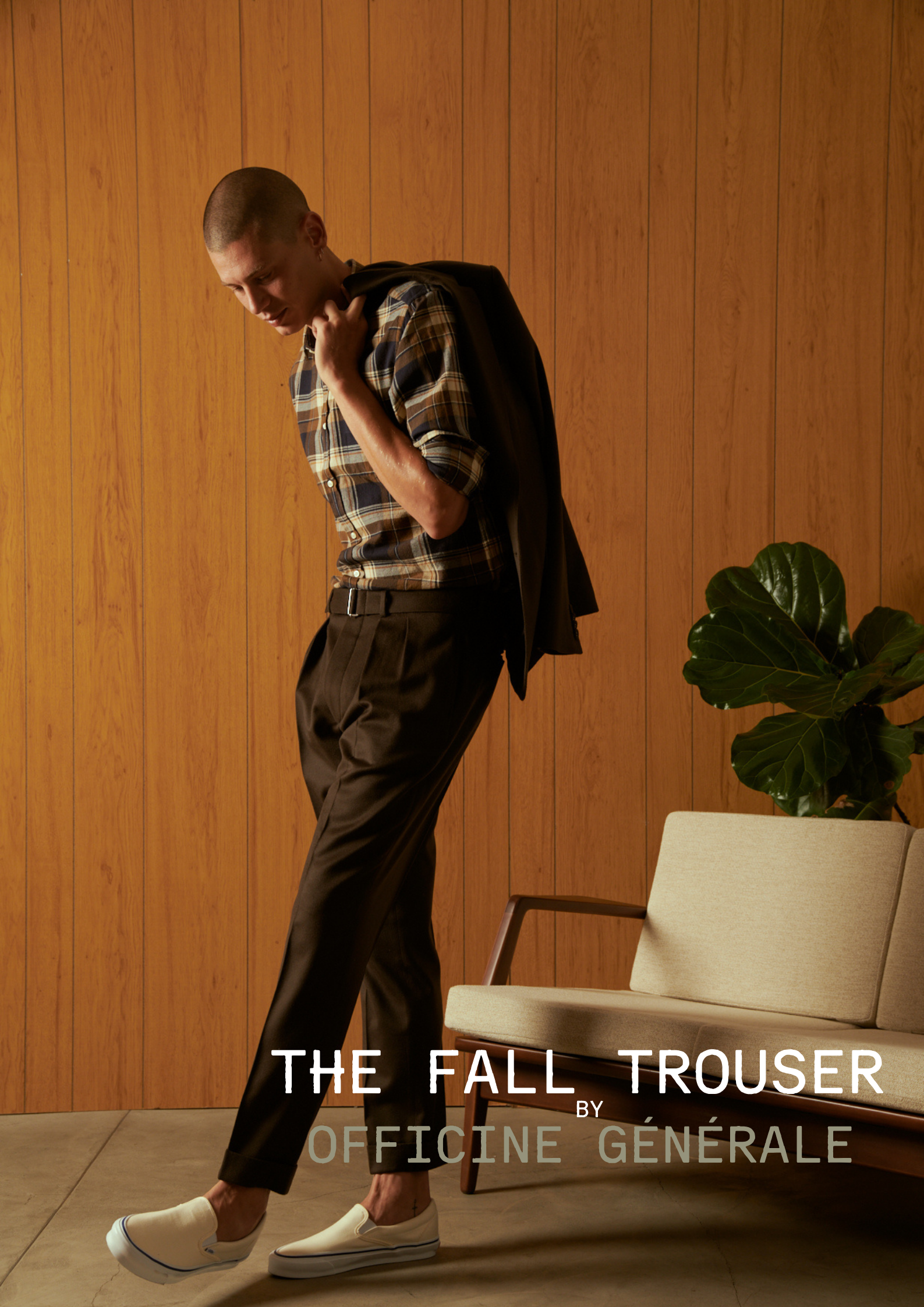 A person in fall trousers and white slip-on shoes stands in an indoor setting with a wood-paneled wall. They are holding a jacket over their shoulder, head tilted downward, with a potted plant and a beige armchair nearby. Text overlay: "The Fall Trou