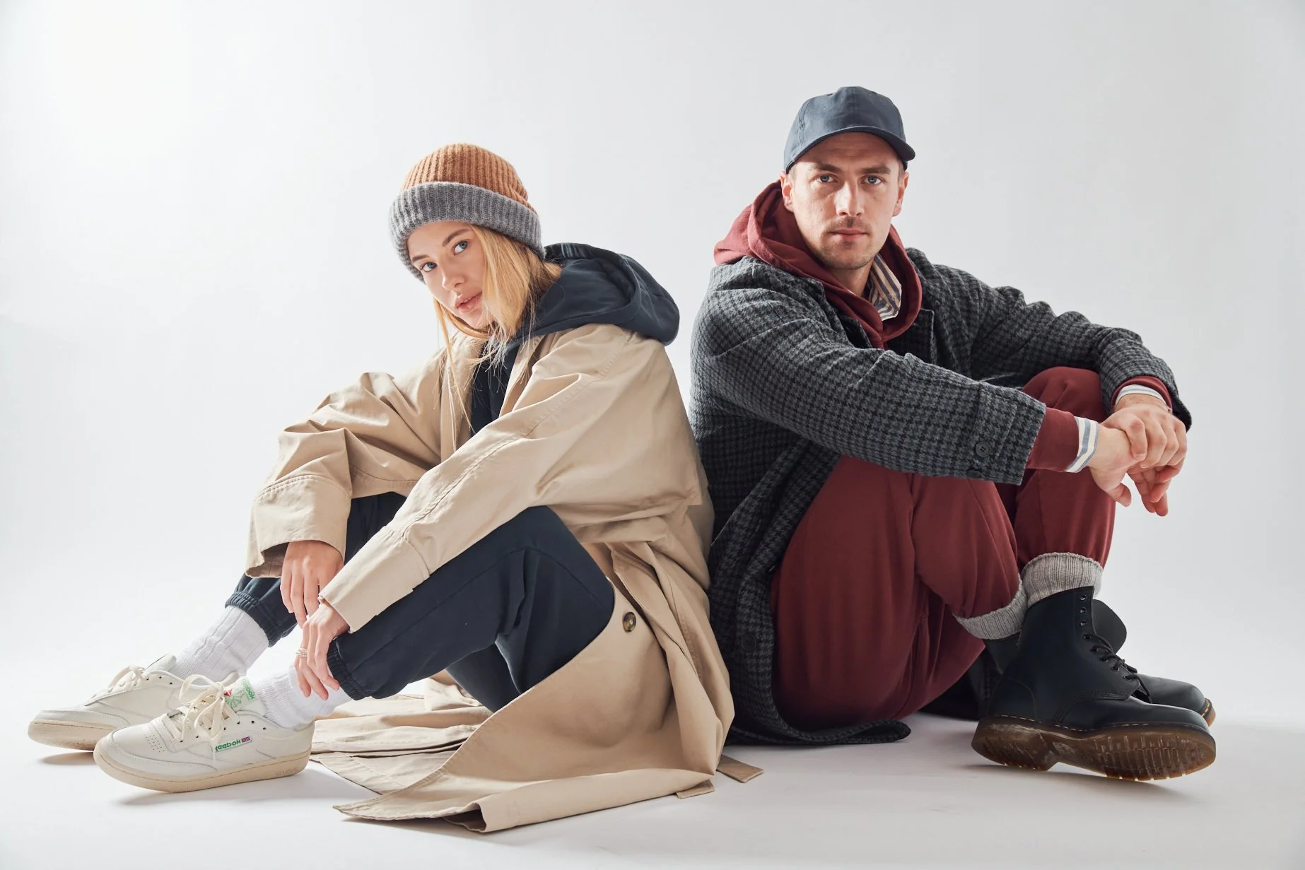 A woman and a man sitting on a white floor against a white background. The woman is wearing a tan oversized jacket, black pants, and white sneakers with Reebok logo, and a brown and gray beanie. Both are sitting with legs bent, leaning slightly to on