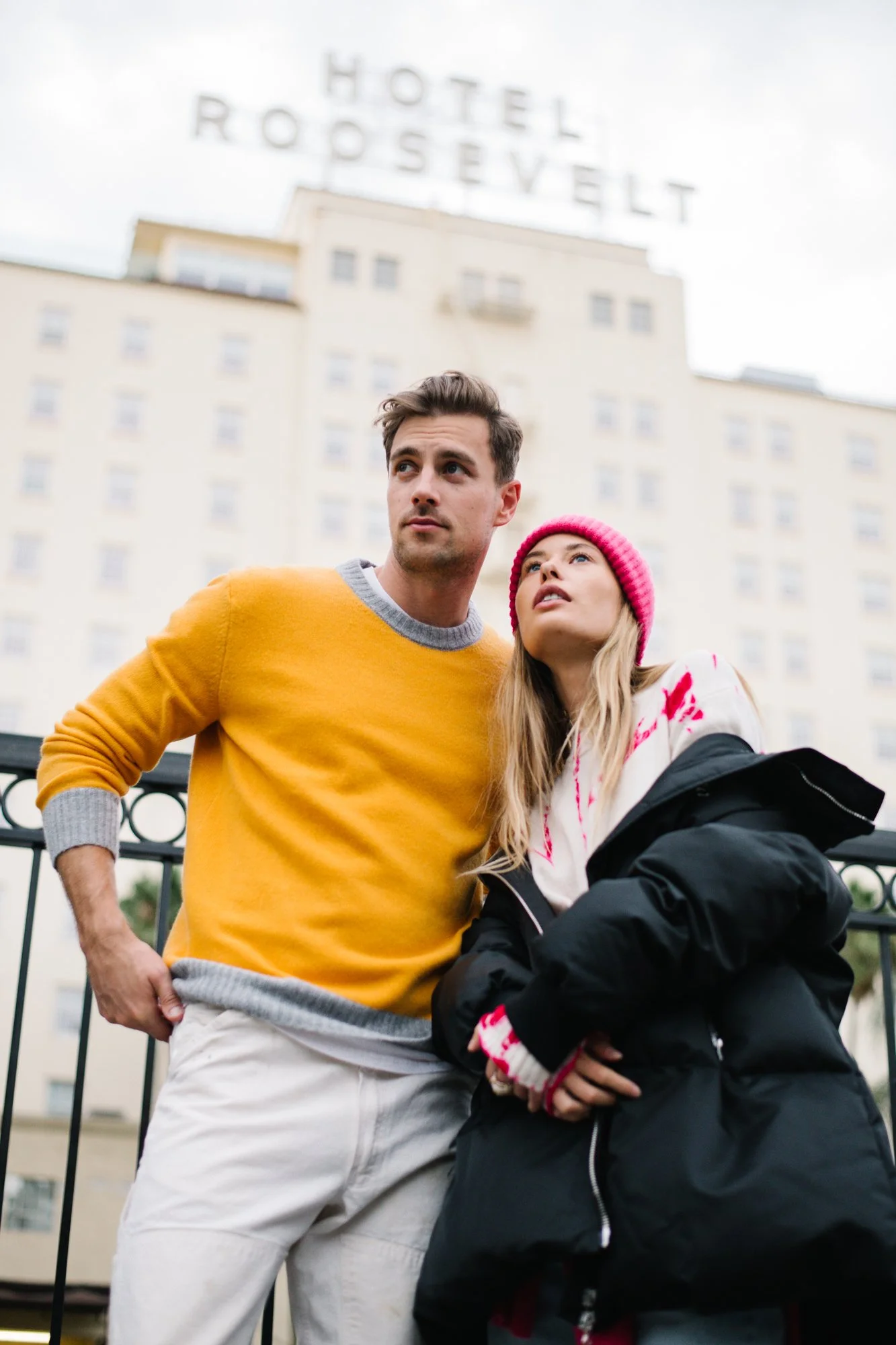 A young man with short dark hair wearing a mustard yellow sweater and a young woman with long blonde hair wearing a pink beanie, a white hoodie with red accents, and a black jacket, are standing close together outdoors in front of a tall building wit