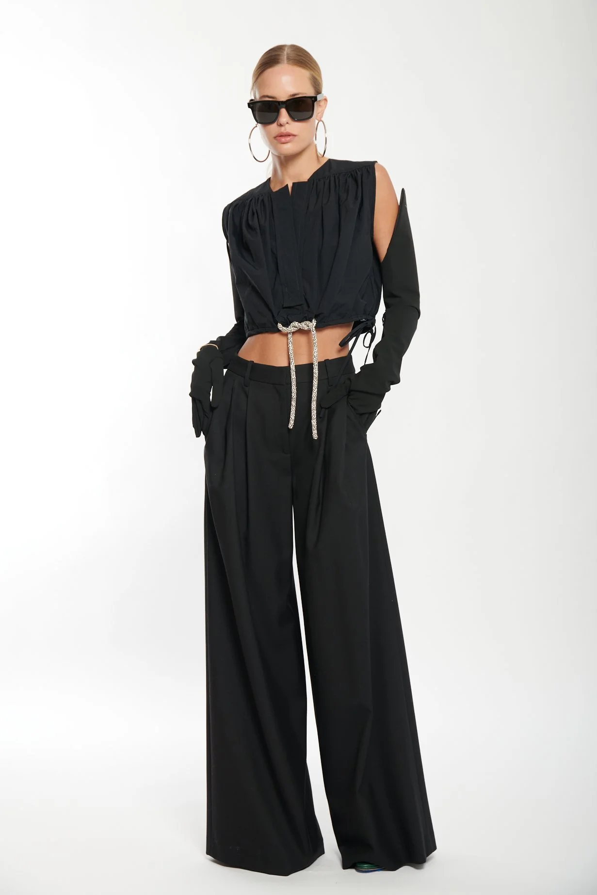 Woman wearing a black crop top, large sunglasses, hoop earrings, black wide-leg pants, and black gloves, standing against a white background.