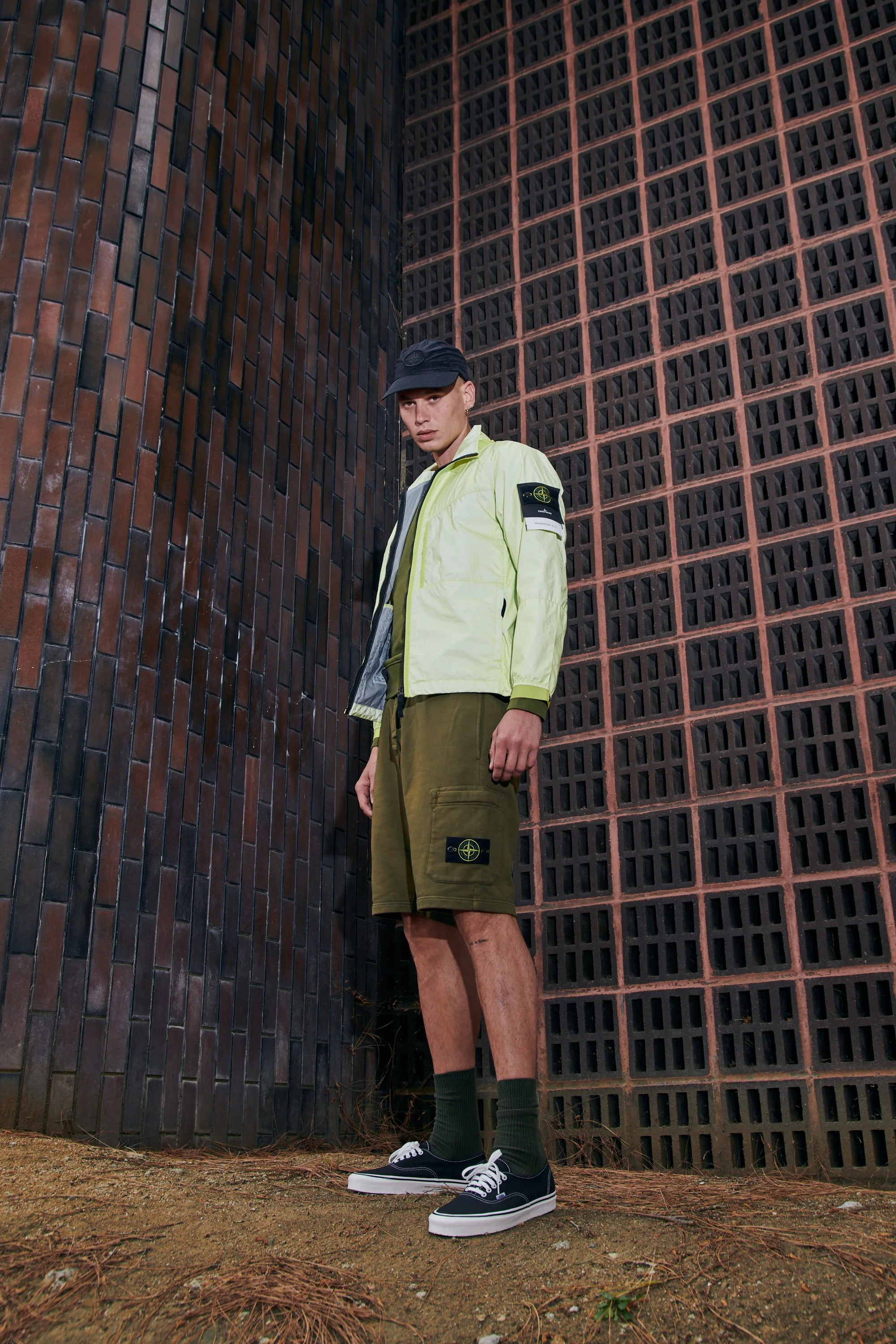 Young man standing outdoors against a brick wall wearing a black cap, a light yellow jacket with a patch on the sleeve, green cargo shorts with a logo, and black sneakers with white soles.