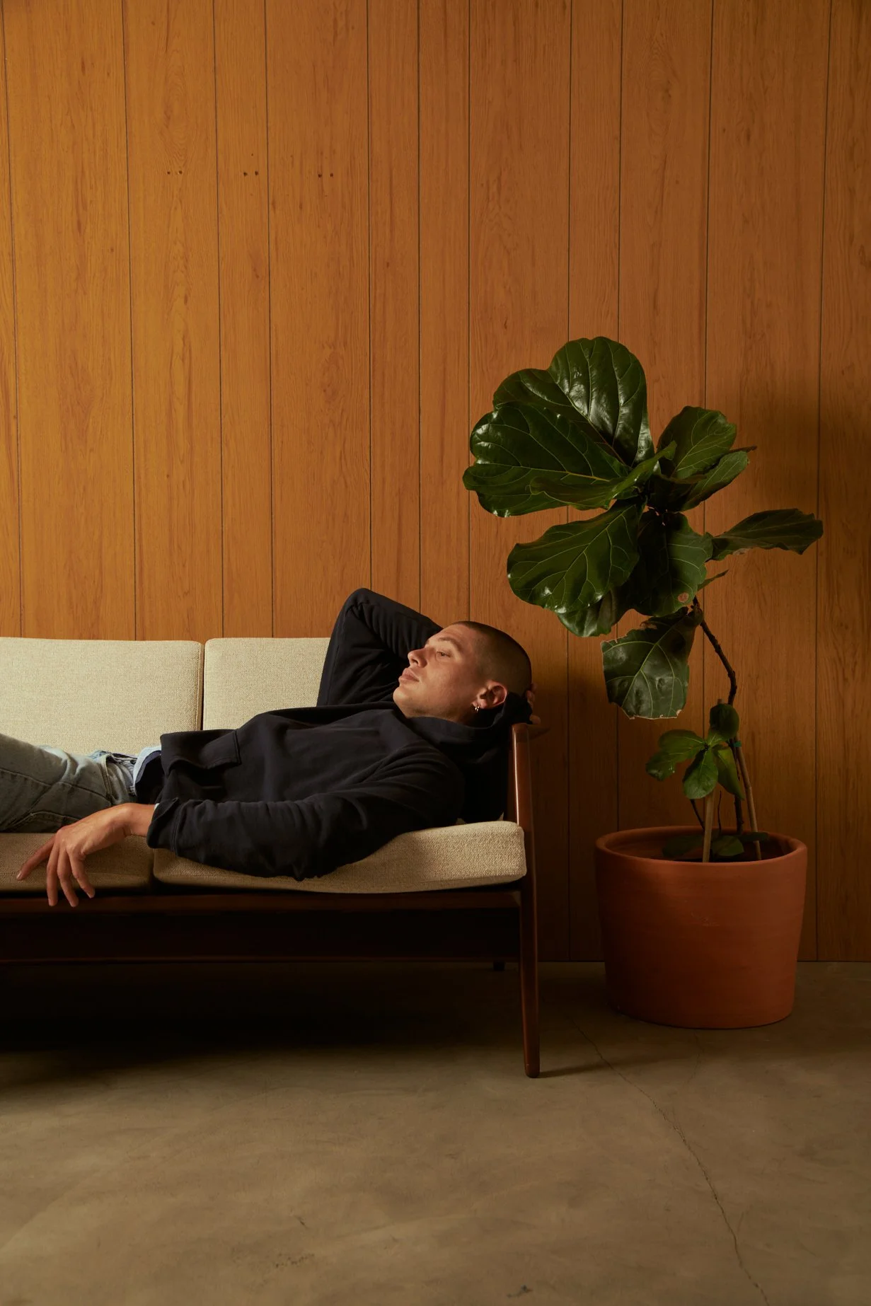 A man with a shaved head and earrings relaxing on a beige sofa with a wooden wall and a large potted fig tree behind him.