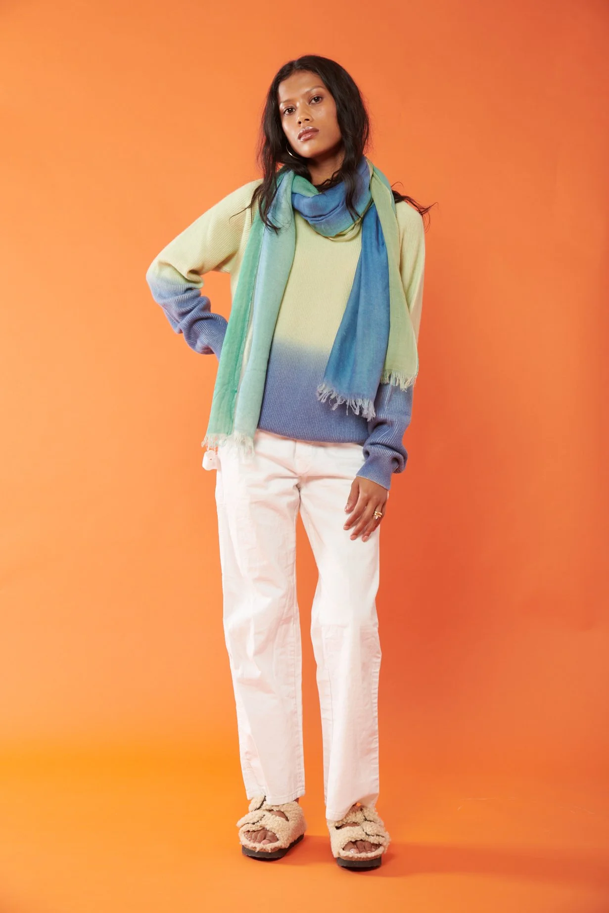 A woman with medium-dark skin tone and long dark hair stands against an orange background. She is wearing a color-gradient sweater, white pants, a multicolored scarf, and fuzzy slippers.
