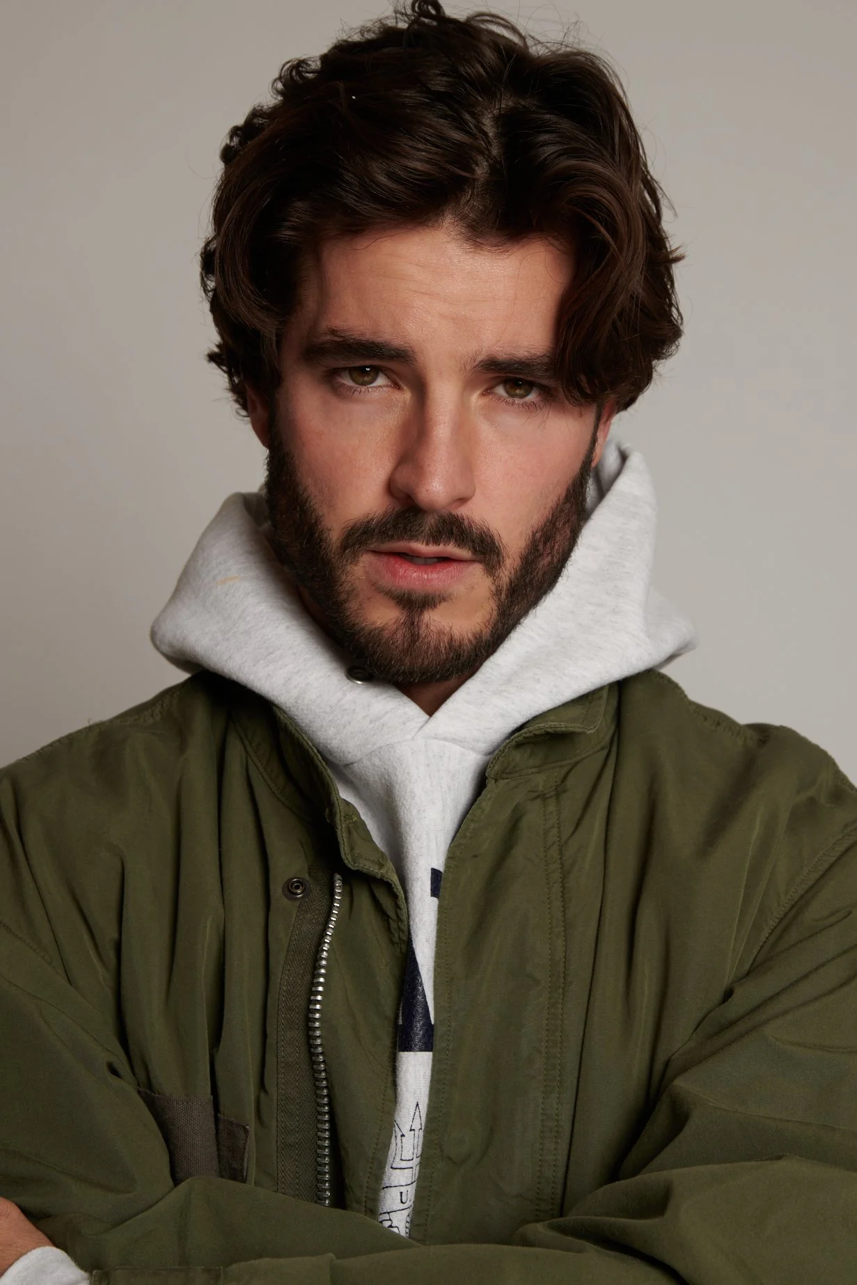 A young man with dark, wavy hair and a beard looking directly at the camera. He is wearing a gray hoodie and a green jacket, with his arms crossed in front of him. The background is plain and light-colored.