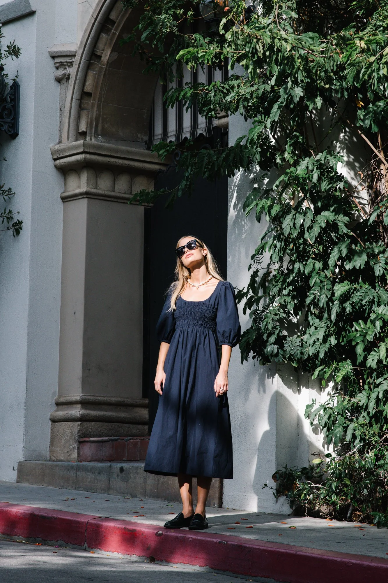 A woman in a long navy dress and black shoes standing on a city sidewalk, wearing sunglasses, with greenery and a building facade in the background.