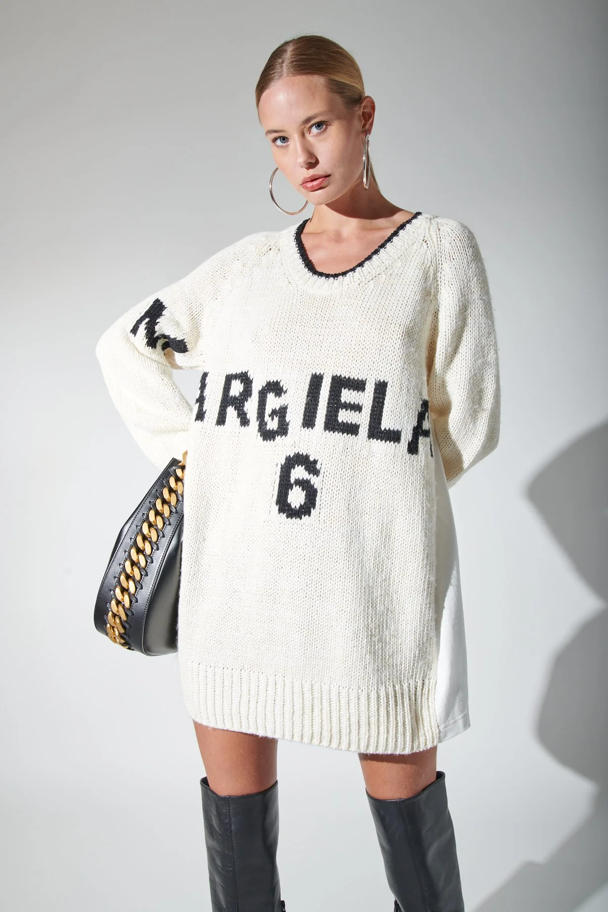 A young woman with blonde hair, wearing large hoop earrings, a white oversized sweater with black lettering that says "MARGIELA 6", a black handbag with gold chain details, and black knee-high boots, standing against a plain background.