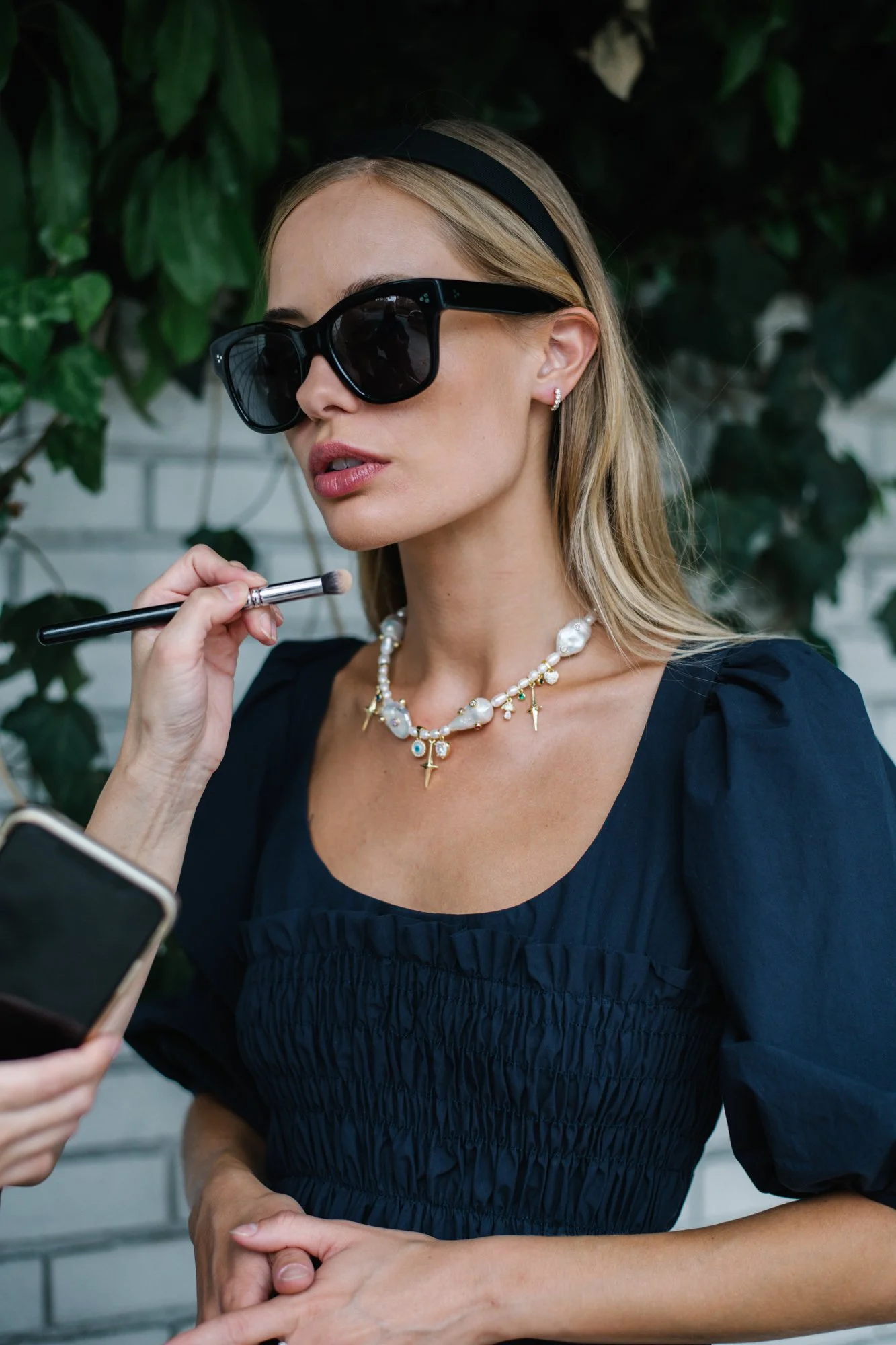 A woman with blonde hair, wearing large black sunglasses, a black headband, and jewelry including a pearl necklace with cross charms, is getting her makeup done with a brush. She is dressed in a black top with puffy sleeves, and her hands are clasped