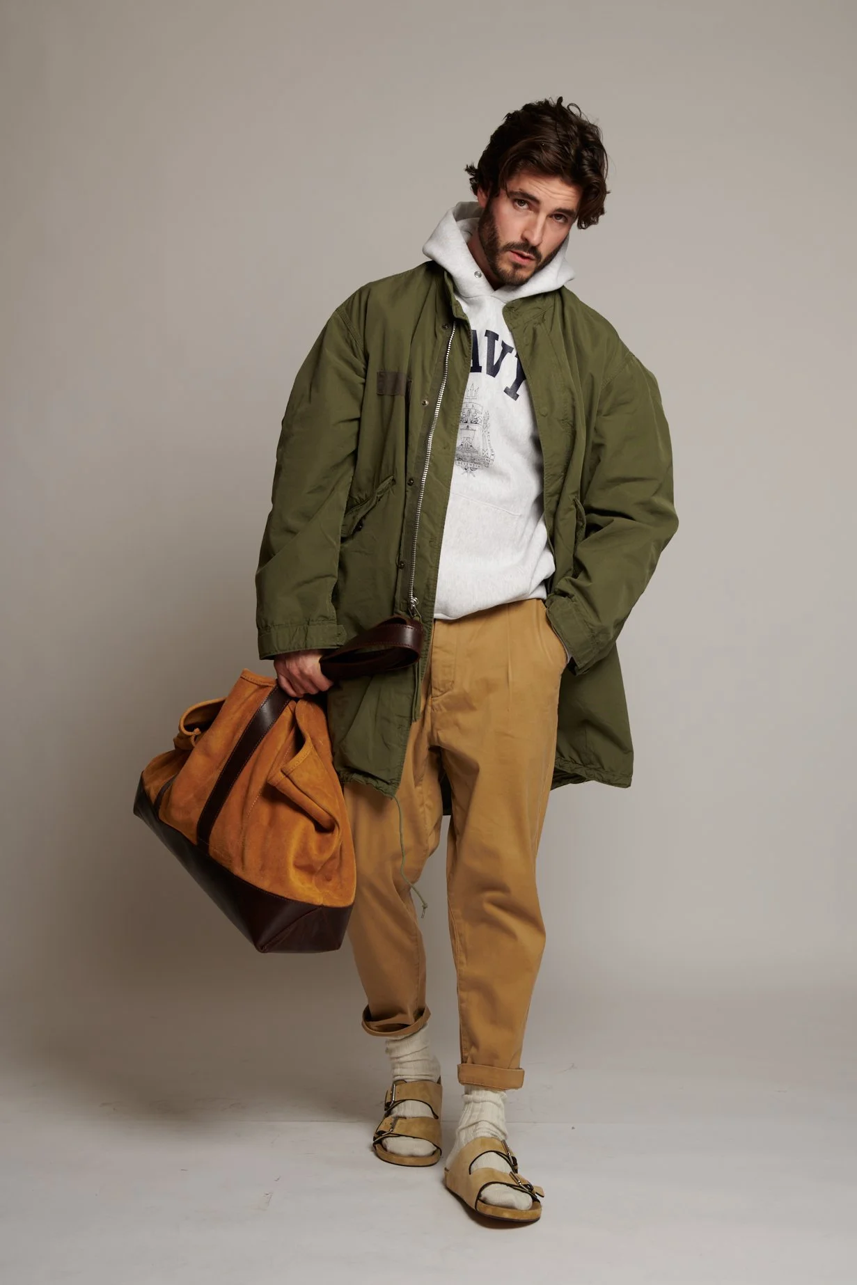 A man with dark, curly hair and a beard posing against a plain gray backdrop. He's dressed in an olive green jacket, a white hoodie with a graphic design, tan pants, and sandals with socks. He is holding a large bag in his right hand and has one hand