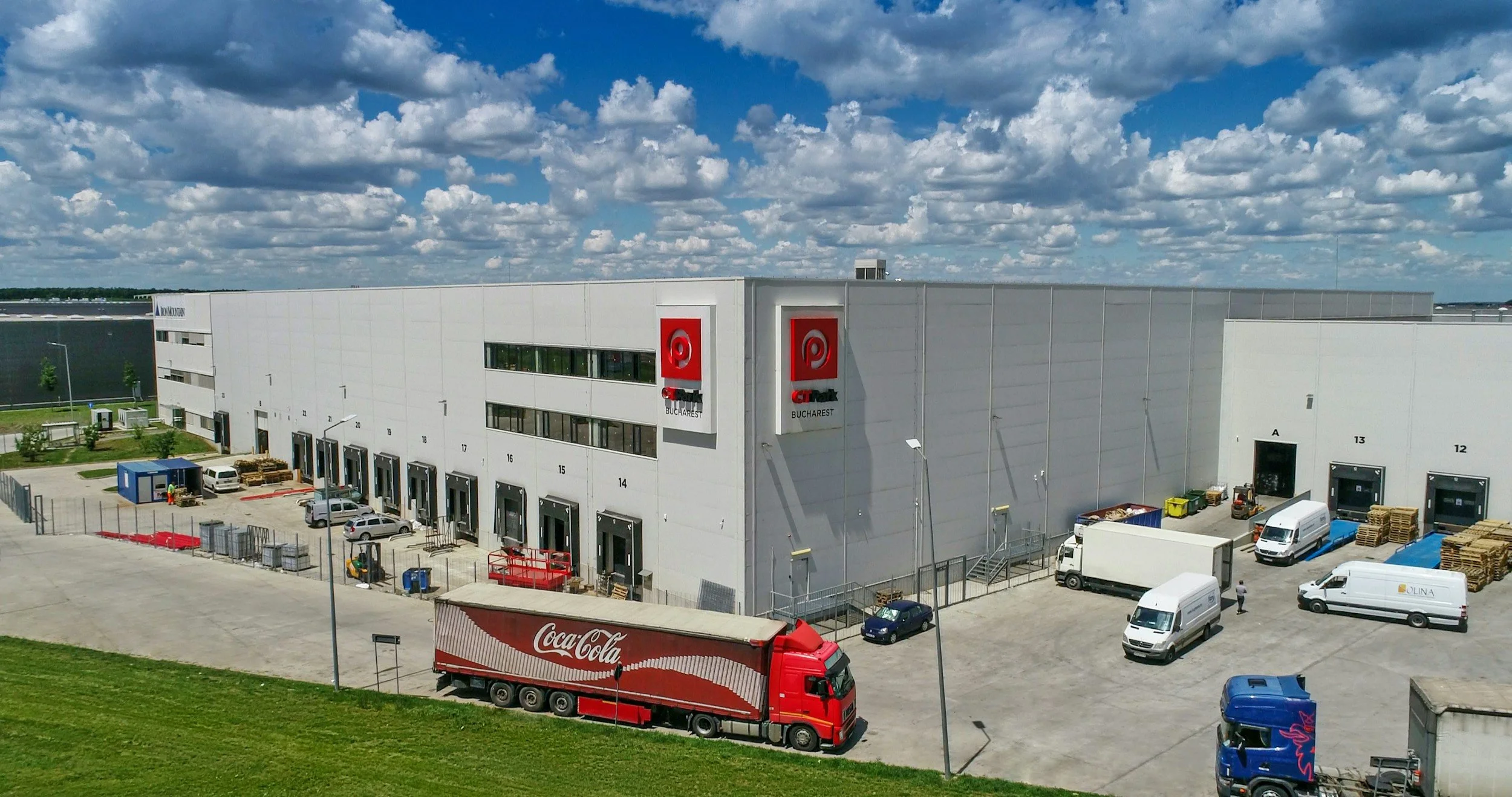 Large white warehouse with multiple loading dock doors, labeled with numbers, and trucks parked outside. The warehouse has signs for 'Coca-Cola,' 'Target,' and 'Lina.' There are trucks and vans nearby, and a partly cloudy sky overhead.
