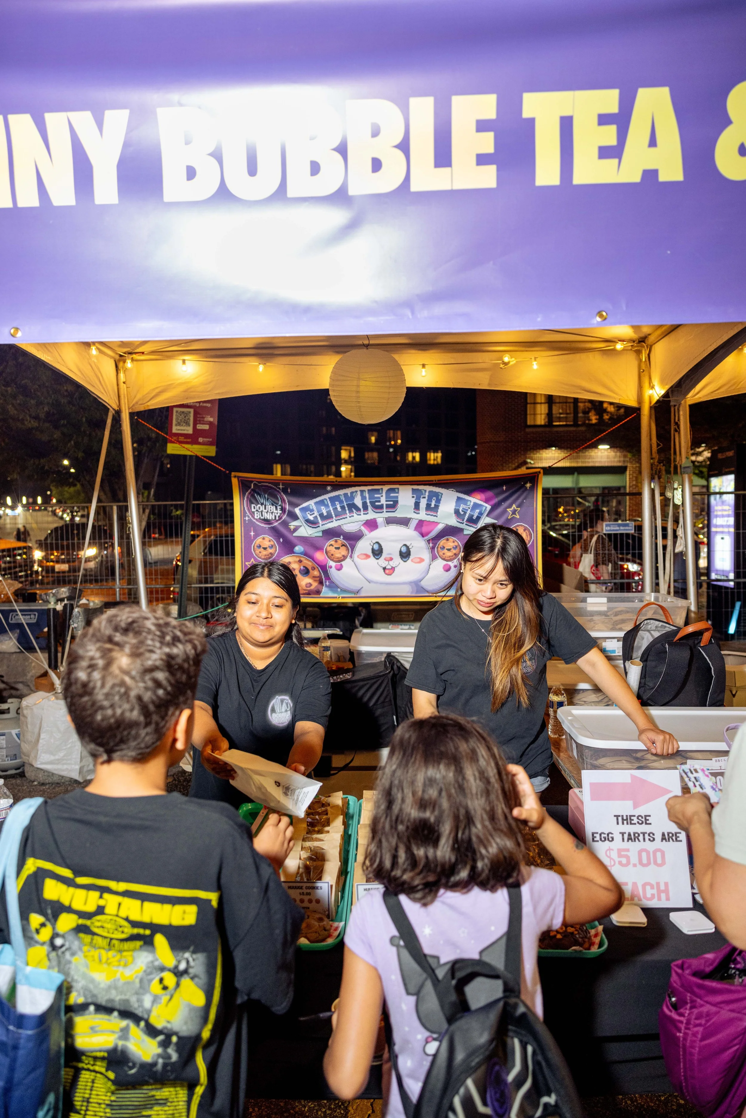 Night scene at an outdoor food stand with people ordering and receiving food, a purple sign overhead reads 'Tiny Bubble Tea', vendor women serving customers, decorated with a poster featuring a cartoon character and cookies.