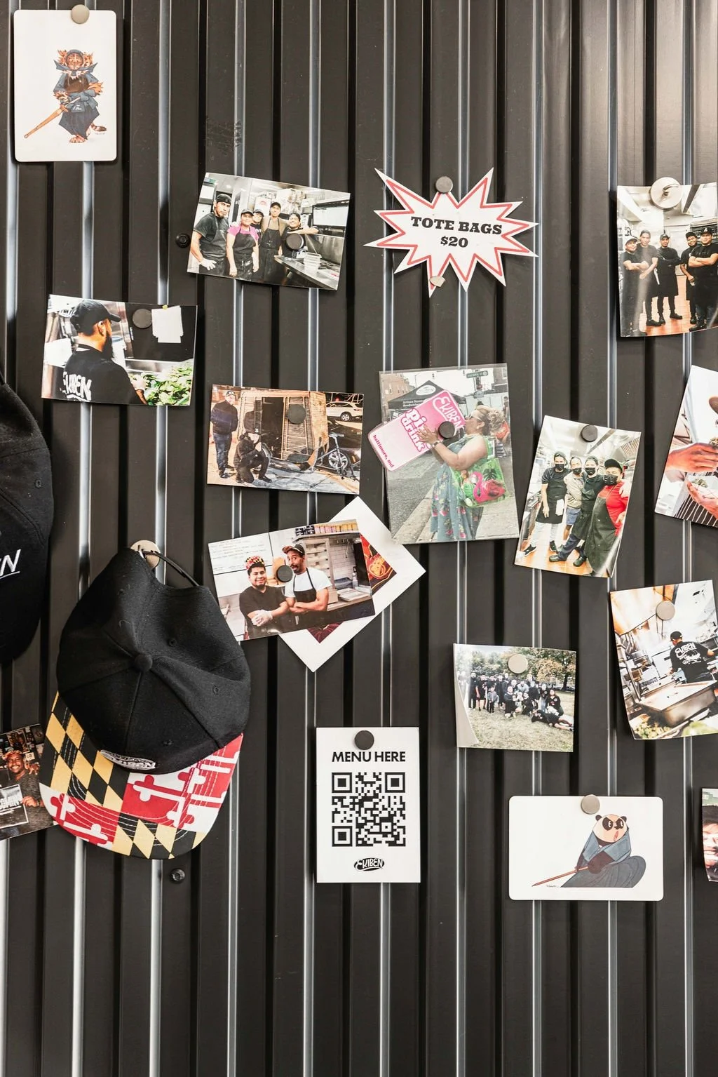 A black metal wall with various printed photographs of people and scenes, a sign that reads 'Tote Bags $20', a QR code for the menu, and a black cap with a multicolored checkered brim hanging from a hook.