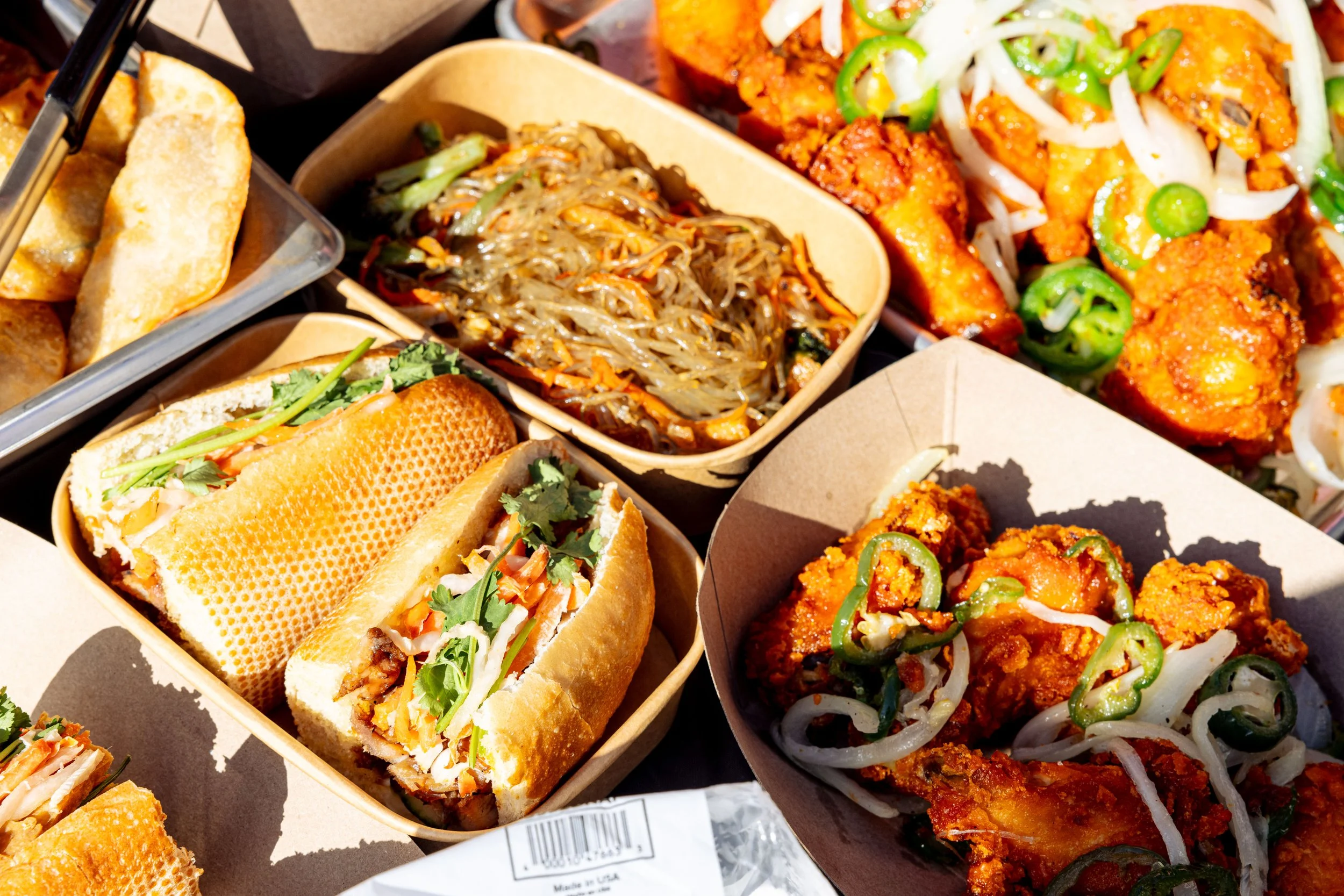 Assorted Asian cuisine dishes in takeout containers, including spring rolls, noodles, and spicy fried chicken topped with sliced green onions and shredded white vegetables.