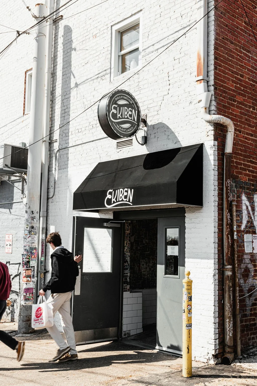 The storefront of a restaurant named 'EkiBen' with a black awning and circular sign, both displaying the name. The building has white and red brick walls, with a person walking past carrying a tote bag.