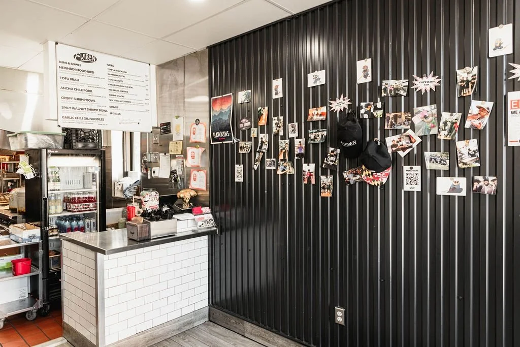 A wall decorated with photos, notes, and bags inside a fast food restaurant or cafe, with a counter and menu board visible on the left.