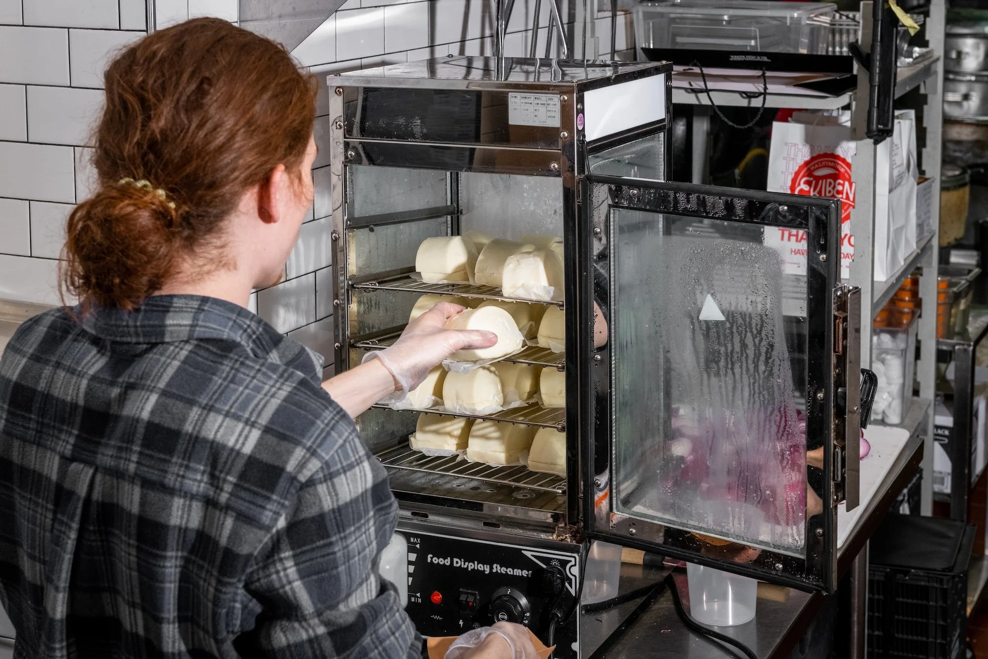 A person with red hair wearing a plaid shirt is placing cheese slices into a food display steamer in a kitchen or food preparation area.