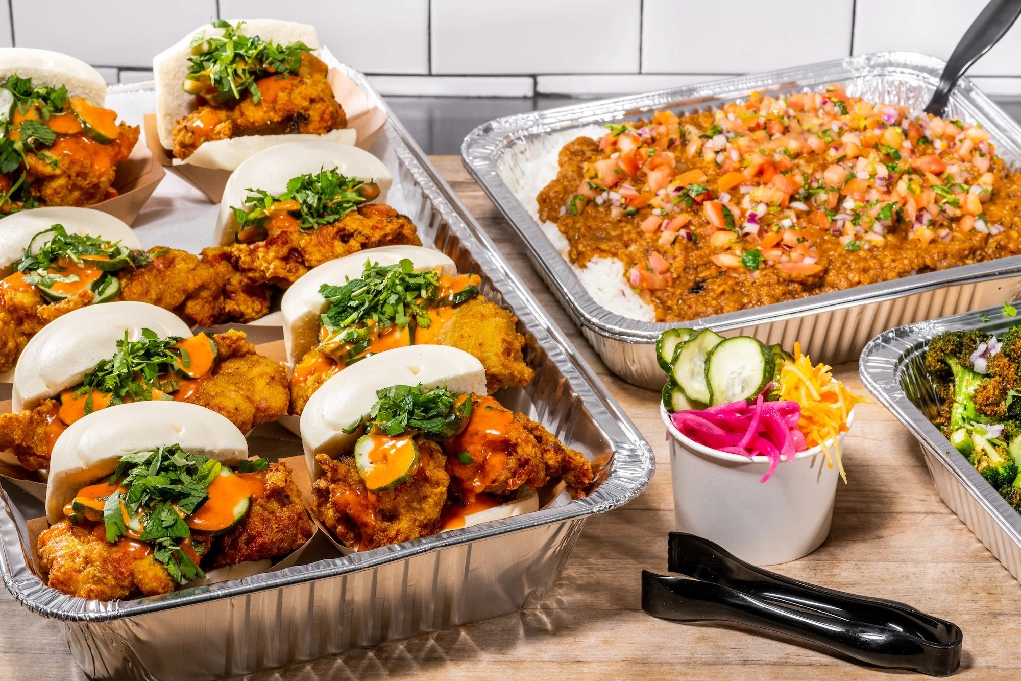 Assorted trays of Mexican food including tacos filled with fried chicken, garnished with cilantro, and white buns, along with a tray of chili con carne topped with diced vegetables, and side dishes of pickled onions, cucumbers, and broccoli. Black tongs are placed on the wooden table.