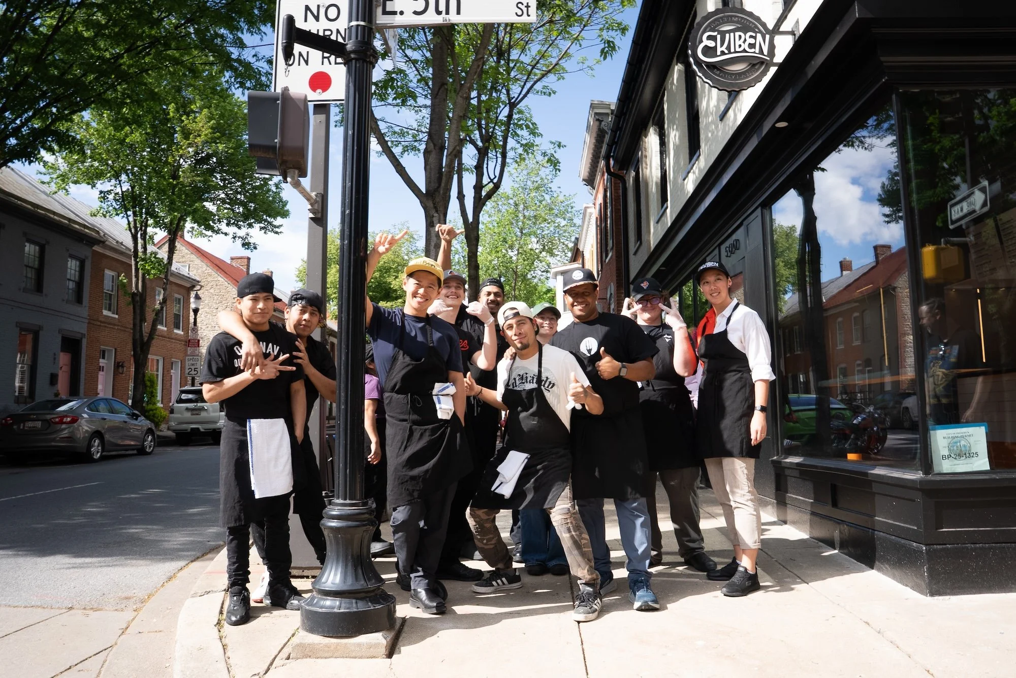 A group of people, including chefs and staff, standing together outdoors on a city sidewalk in front of a storefront with a sign that reads 'E. 5th'. They are smiling and posing for the photo, with some making gestures. The street behind them has parked cars and trees, and there is a street sign overhead.