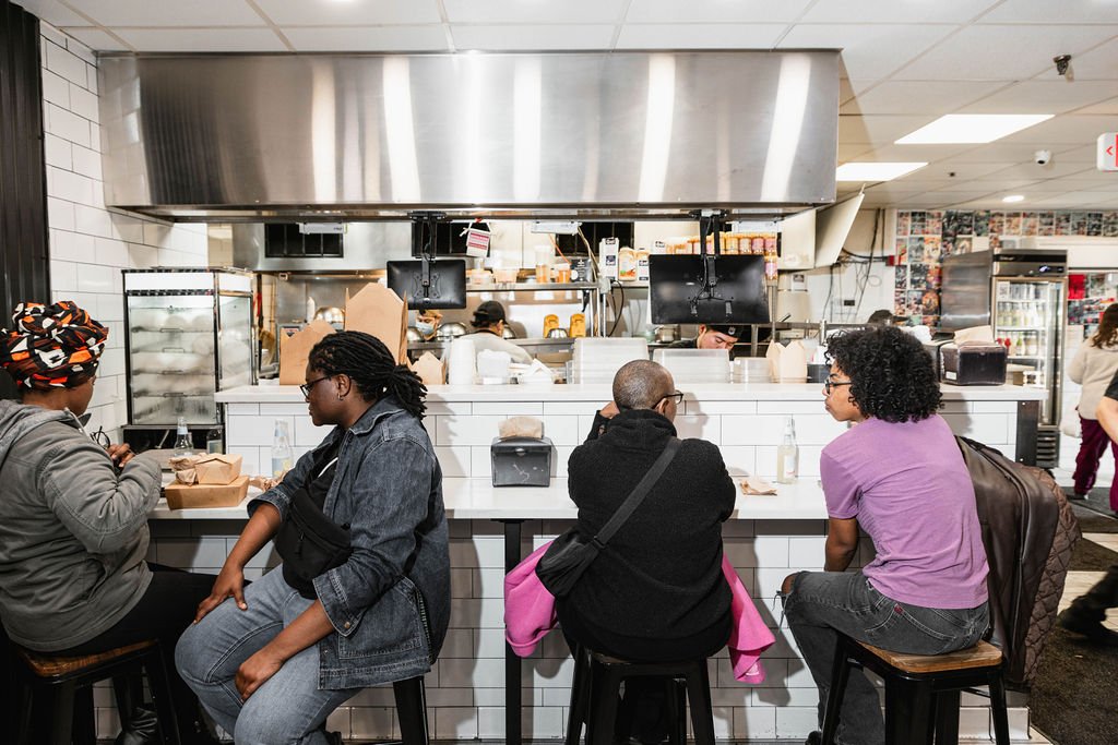 Four women sitting at a counter in a restaurant or cafe, with the kitchen visible behind them, as they have food and drinks.