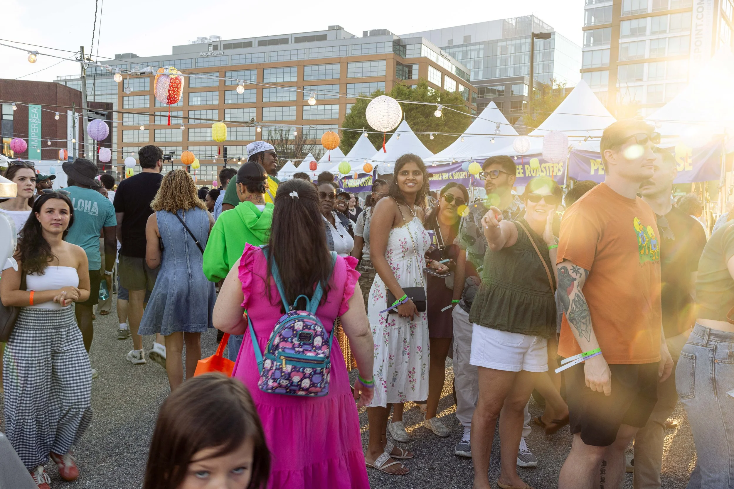 Crowd of people at an outdoor festival or event with colorful lanterns and tents in the background, sunlight glare.