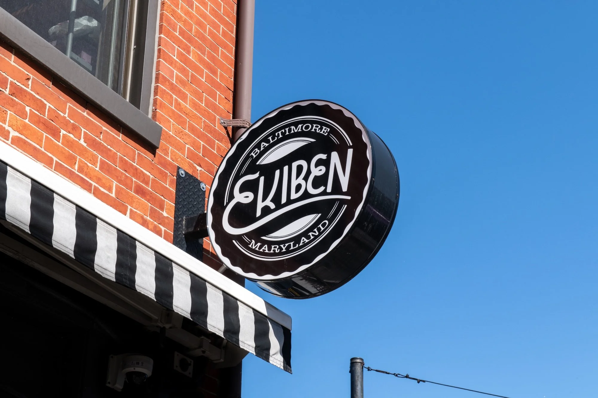 A round black and white sign hanging outside a building reads "Baltimore SKIBEN Maryland" with a skateboard graphic in the design. The building has red brick walls, a window, and a black-and-white striped awning beneath the sign. The sky is clear and blue.