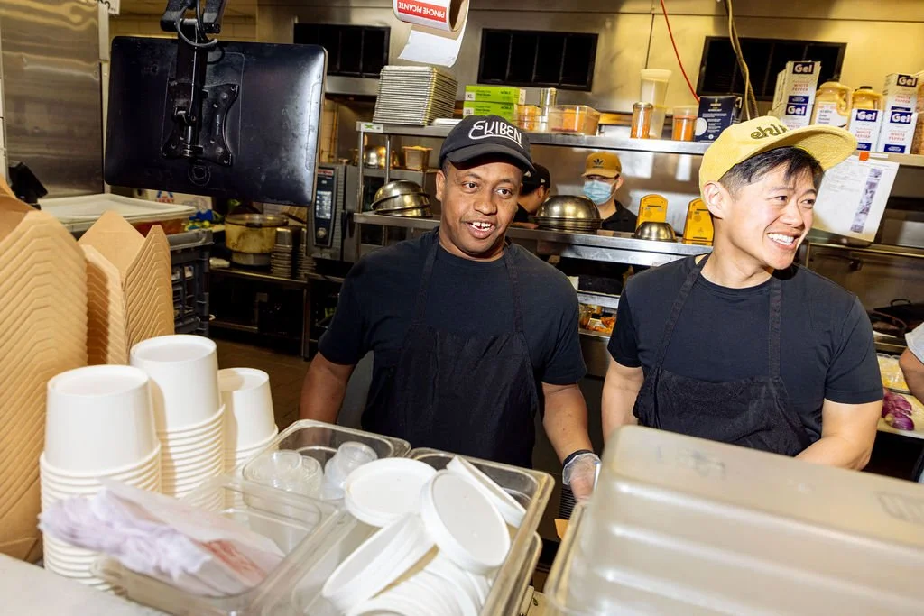 Two male workers behind a restaurant counter, smiling and wearing black t-shirts and aprons. One wears a cap with 'EKIBEN' on it and the other a yellow hat with 'eHe' on it. In the background, shelves with condiments, a person with a mask, and kitchen equipment are visible.