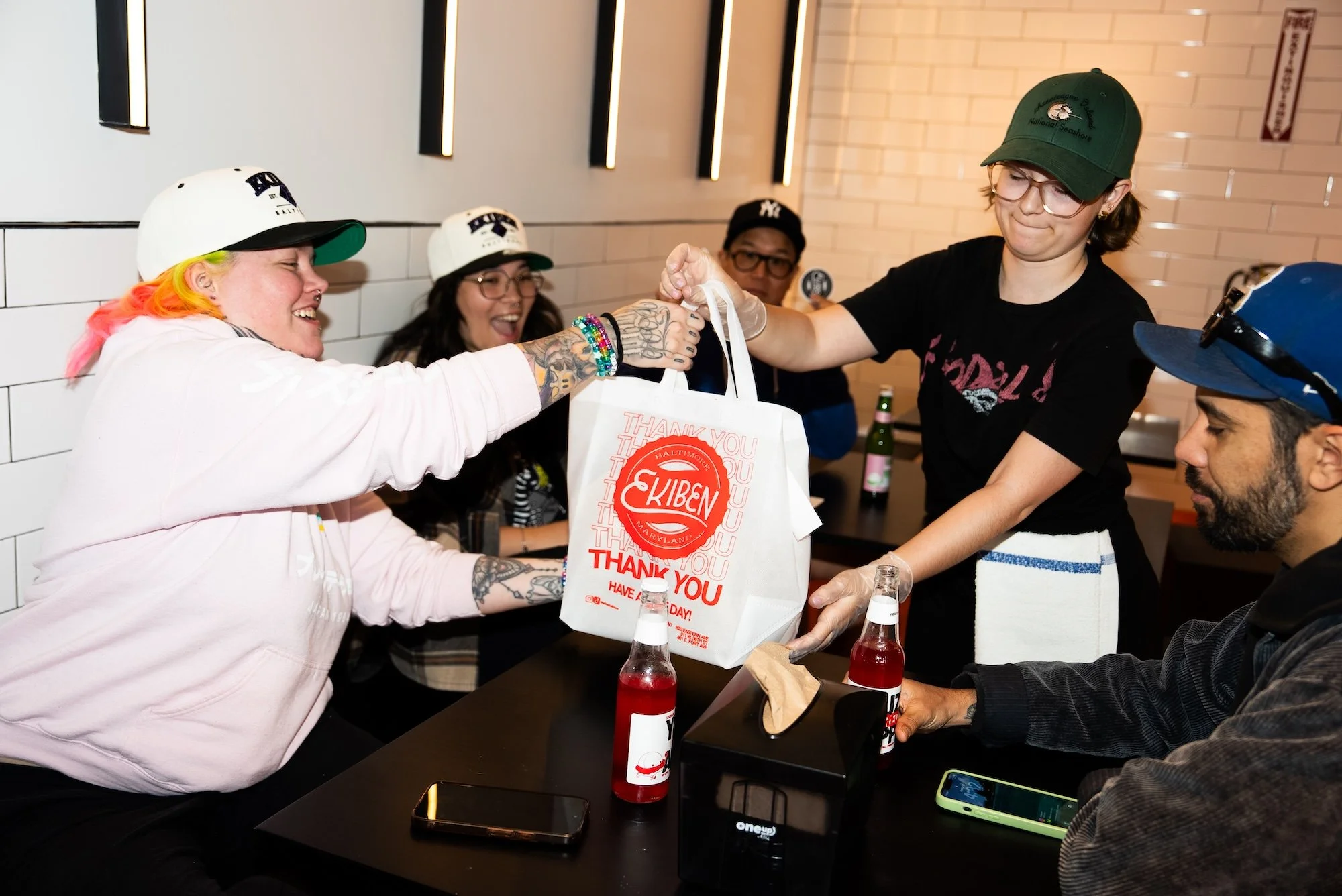 A group of people sitting around a table, with some wearing caps and glasses, exchanging gifts and smiling. One person is handing a white bag with red and white text to another. Bottles of red drinks are on the table. The setting appears to be a casual gathering or celebration.